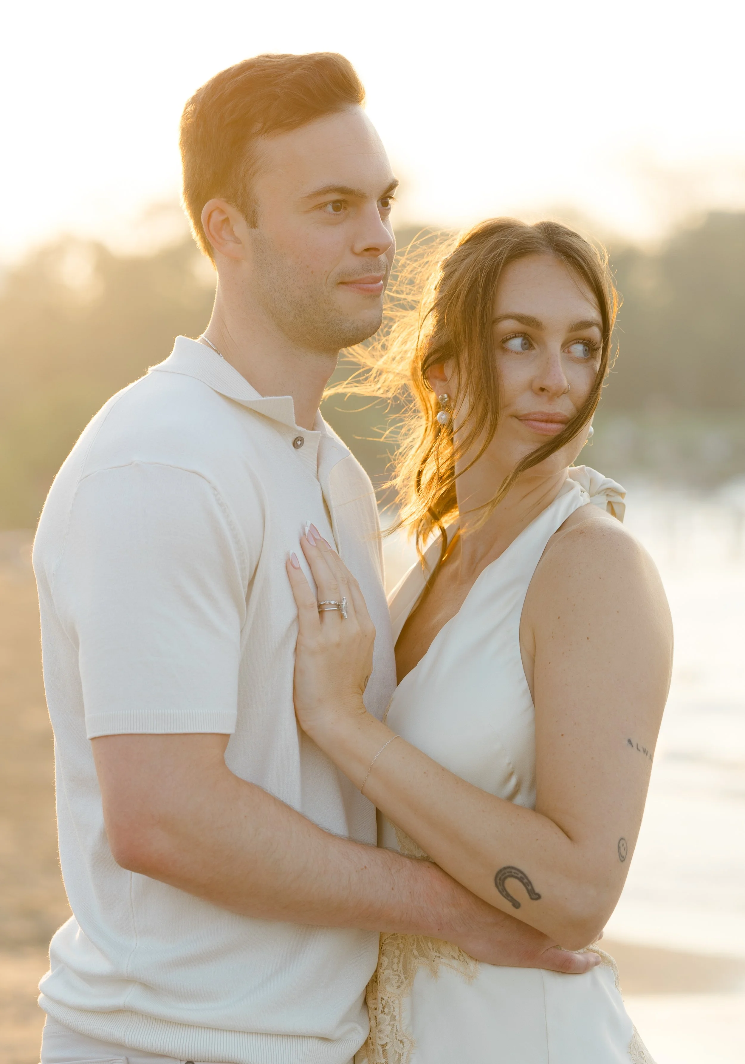 A couple standing closely outdoors at sunset, with the woman's hand on the man's chest, both looking to the side. The man has red hair and is wearing a white polo shirt. The woman has brown hair, blue eyes, tattoos, and is wearing earrings and a whit