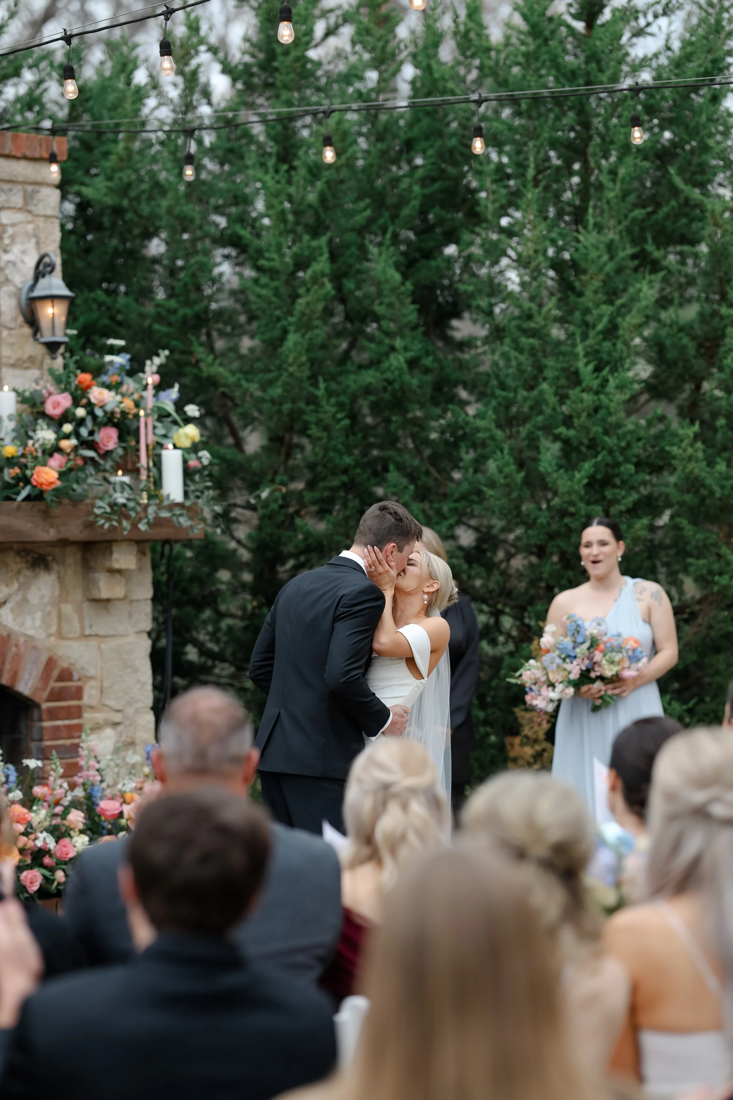A couple kissing during a wedding ceremony outdoors, with guests seated watching, surrounded by green trees, string lights, and floral decorations.