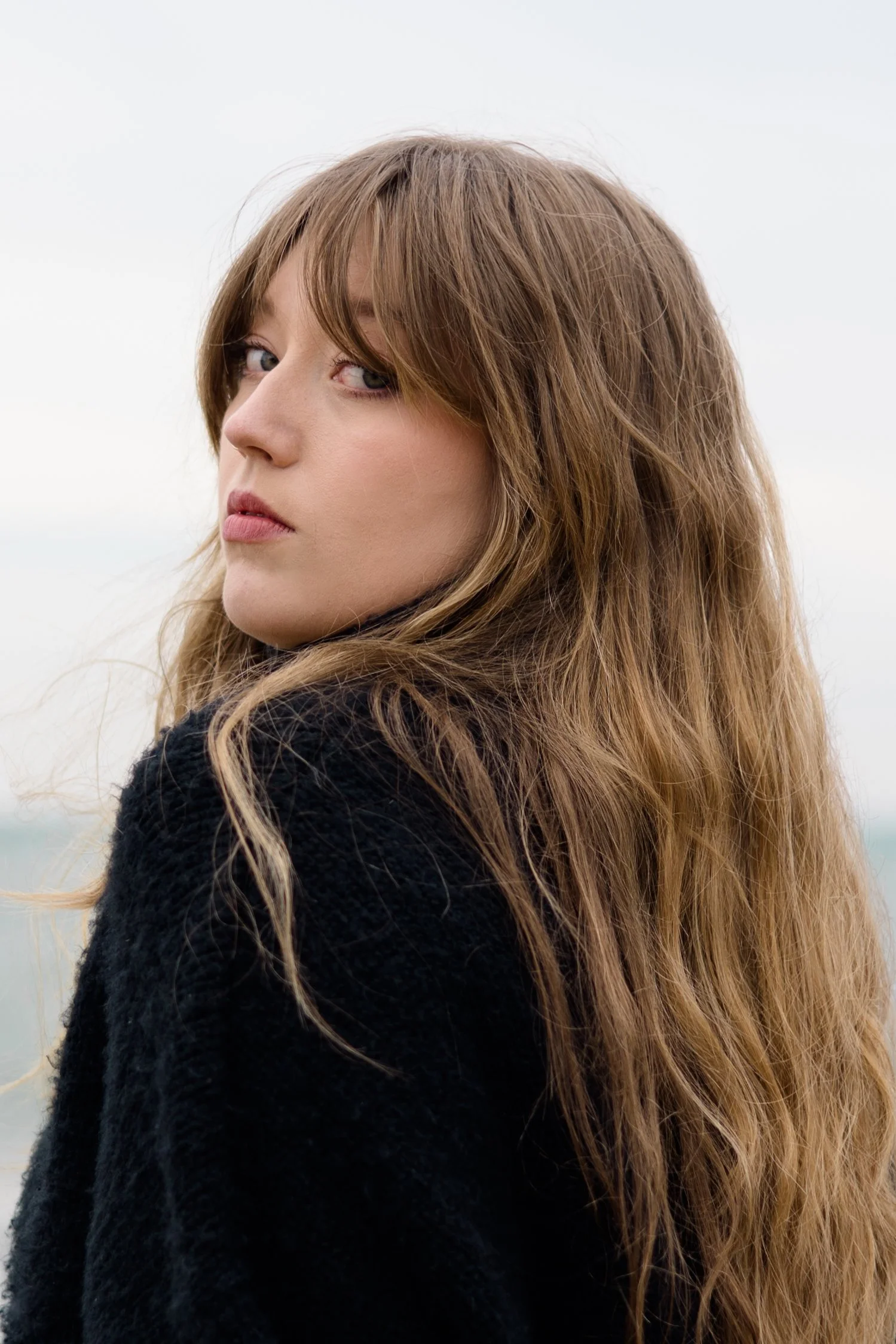 Close-up of a woman with long, wavy, light brown hair and light skin, looking over her shoulder against a pale, overcast sky.