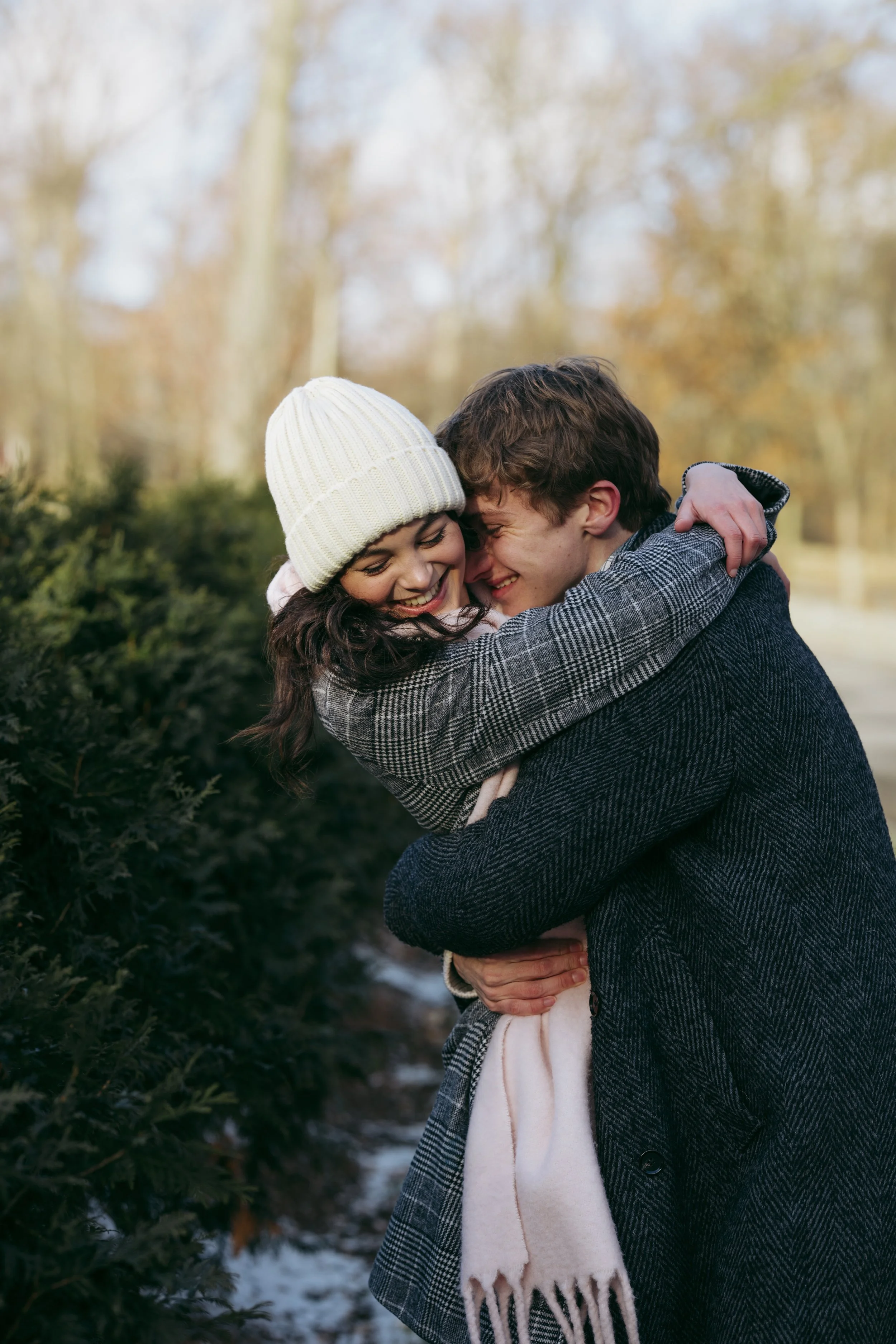 A young couple is hugging outdoors during winter, both smiling and with their faces close together. The woman wears a white knit beanie and the man wears a checkered coat, with a beige scarf around her neck. The background shows blurred trees and bus