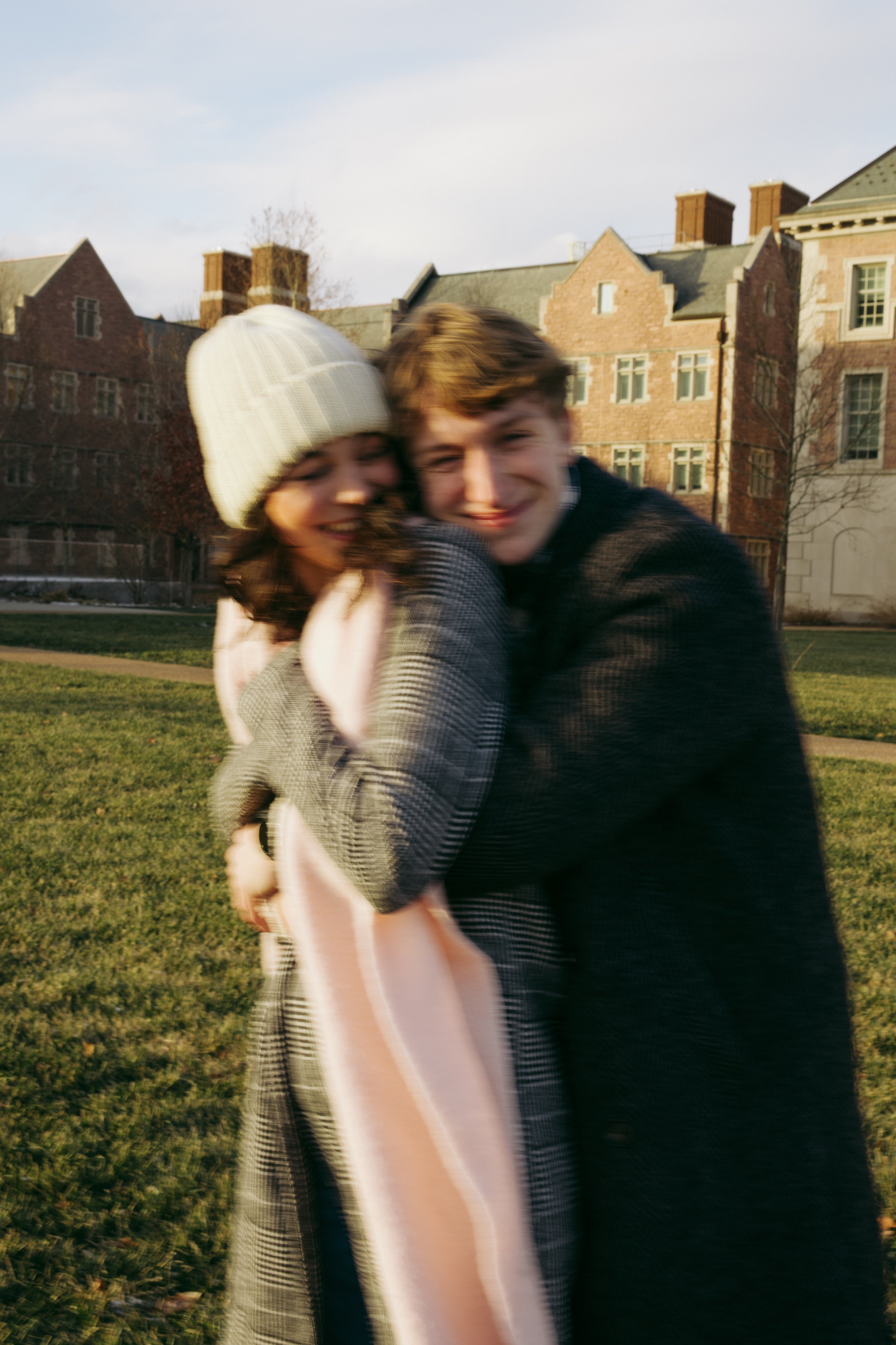 Two young people embracing outdoors with brick buildings in the background, wearing casual clothing and one in a white knit hat, during daytime with cloudy sky.