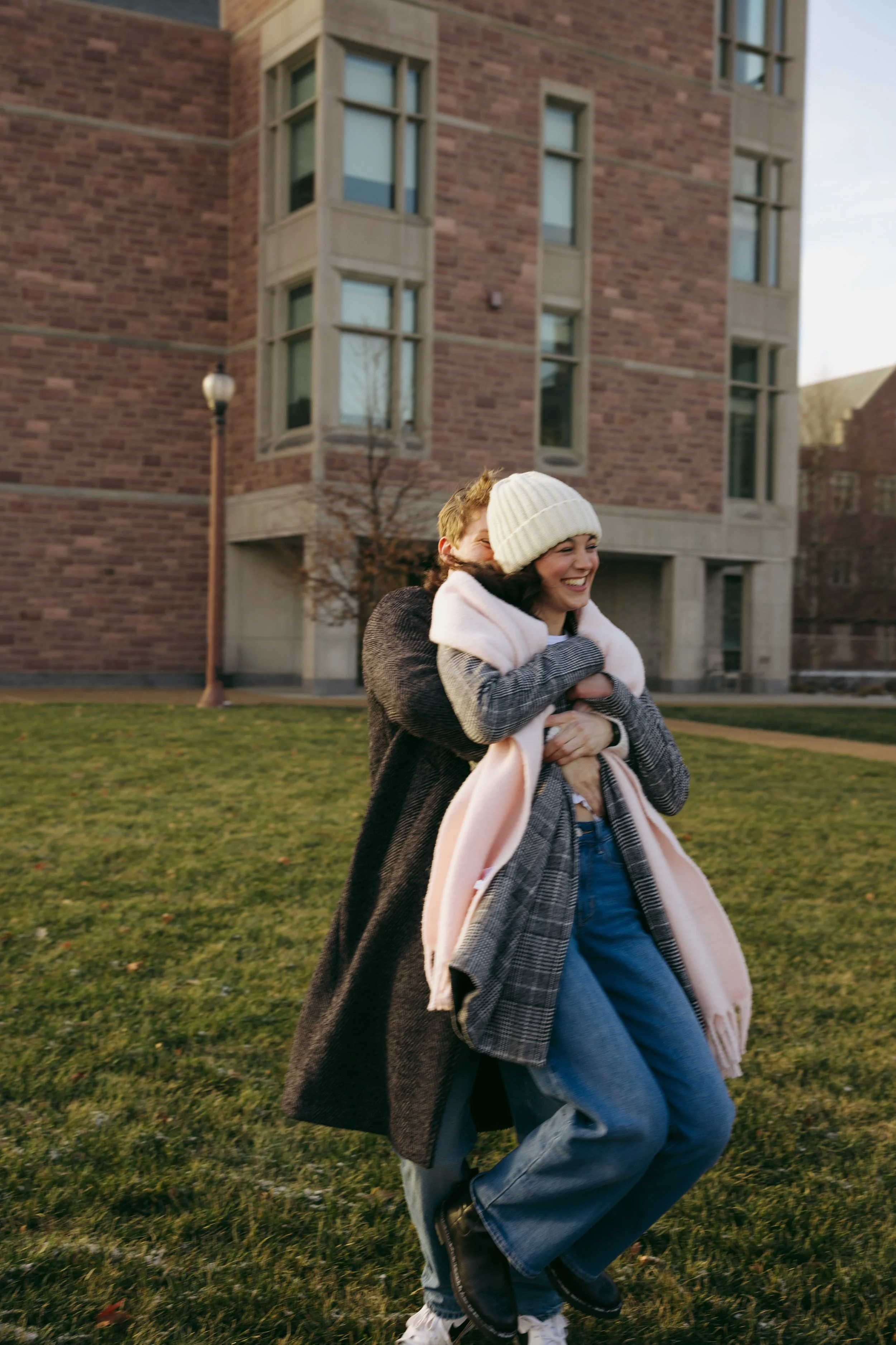 Two people, a man and a woman, are outdoors on a grassy area in front of a brick building. The woman is smiling and wearing a white knit hat, pink scarf, and a coat, while the man is hugging her from behind.