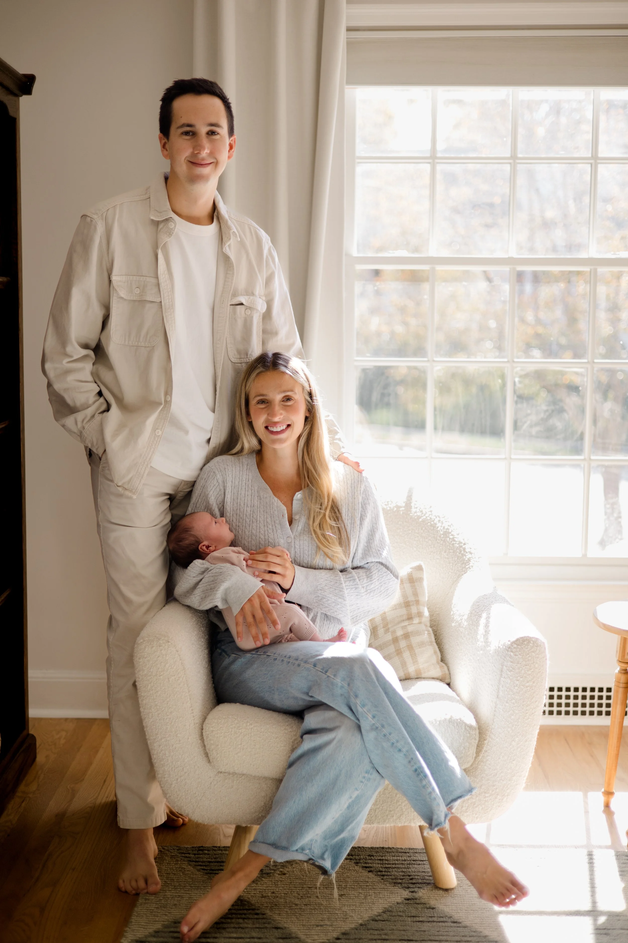 A family of four, including a man, woman, and two babies, poses near a large window in a cozy living room with natural light and neutral-colored furniture.