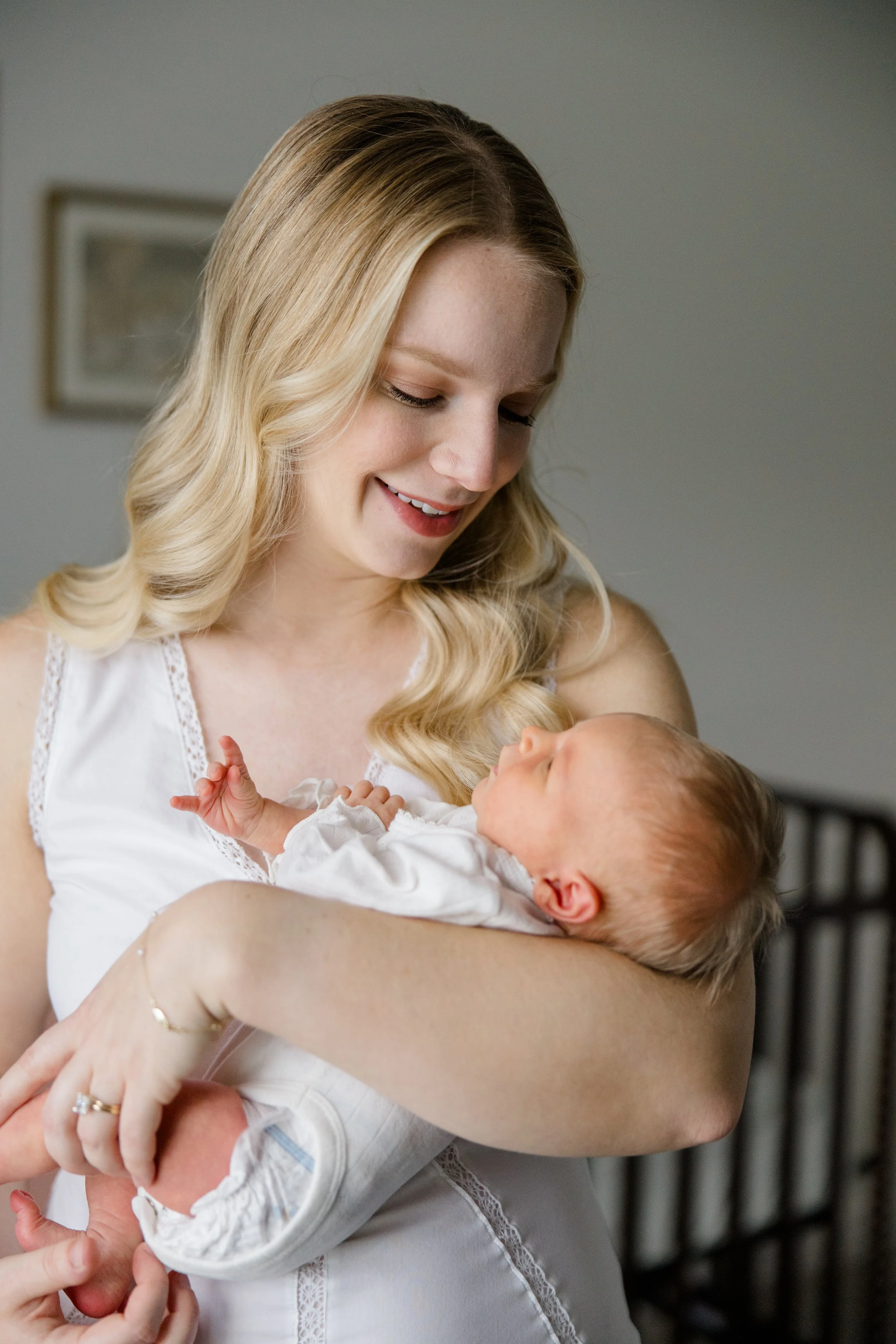 A woman with long blonde hair smiling while holding a newborn baby in her arms, both dressed in white, inside a well-lit room.