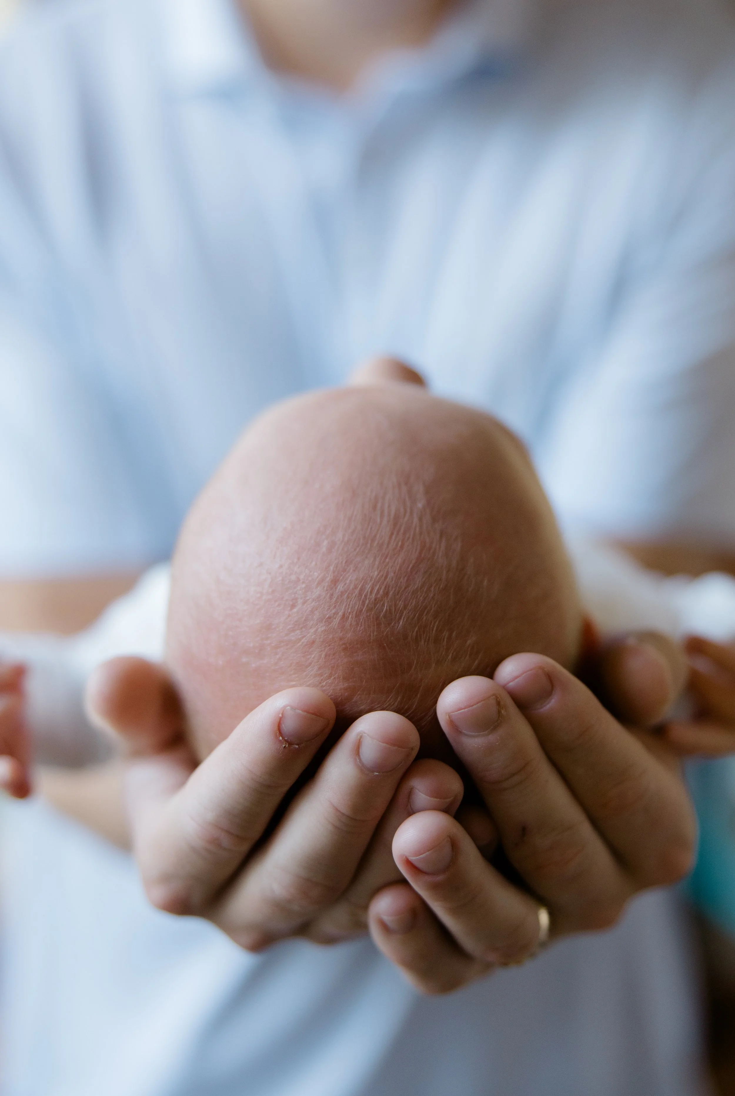 Close-up of a person’s hand holding a reclining baby’s head, which has soft, light skin and very little hair. The person is wearing a ring on their finger, and the background is blurry with soft, neutral colors.