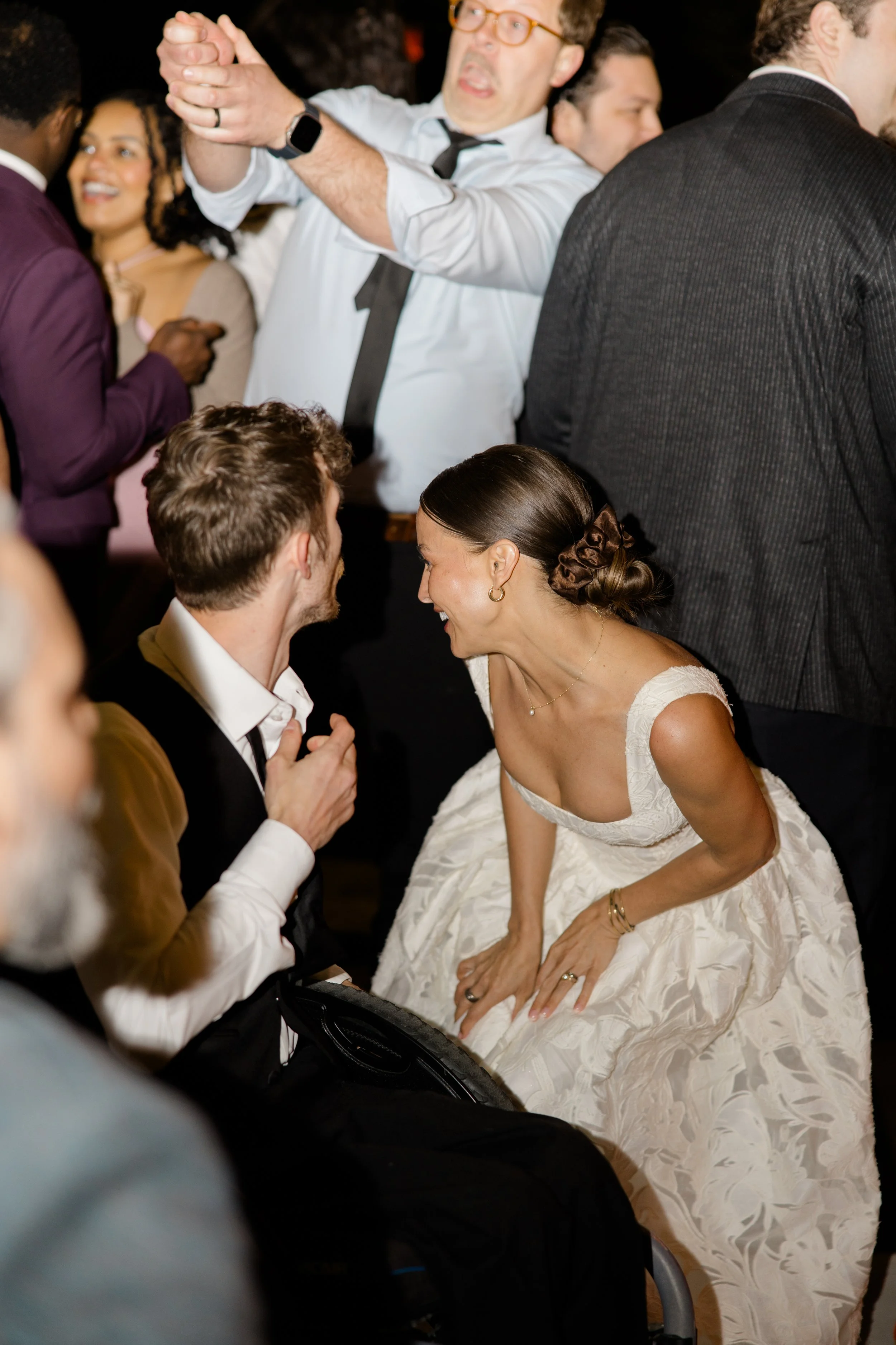 A wedding celebration scene with people dancing and smiling. A bride in a white dress with jewelry is interacting with a man in a suit. Other guests in formal attire are visible in the background, enjoying the event.