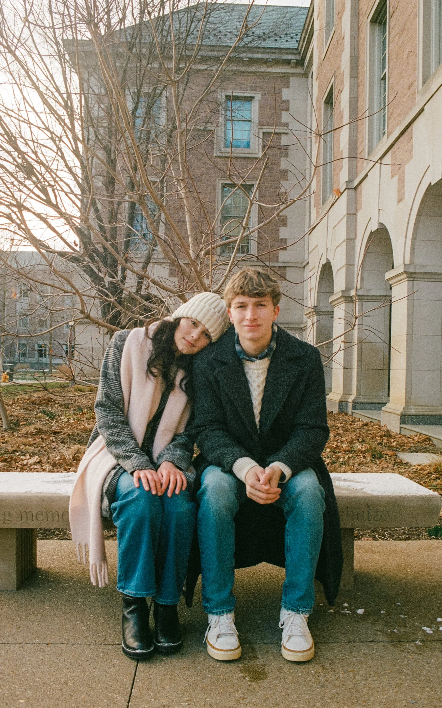 A young woman with long dark hair wearing a beige knit hat and a light pink coat sits beside a young man with short light brown hair wearing a dark coat and light-colored sneakers on a stone bench outdoors. Behind them is a leafless tree and a brick 