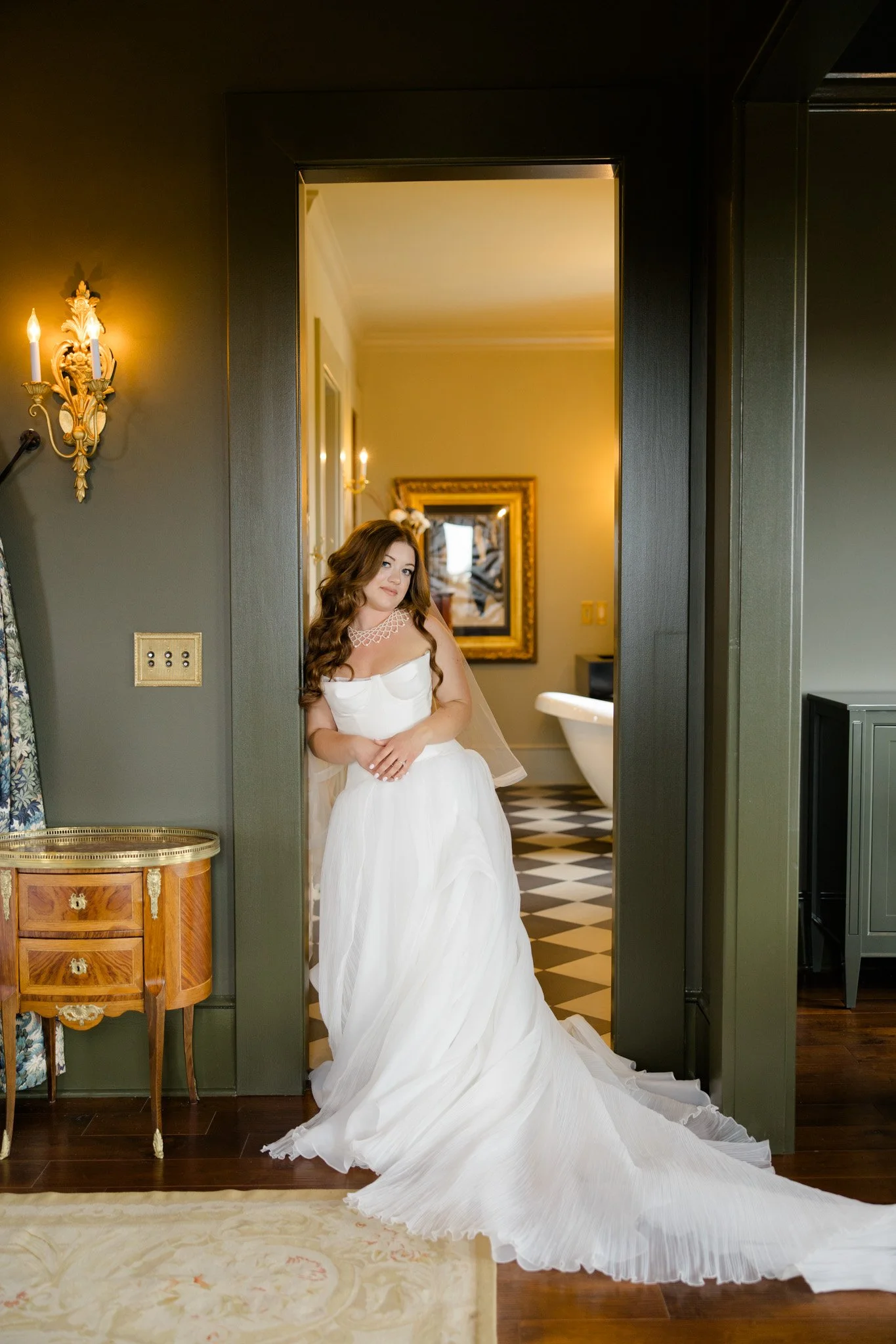 A woman in a white wedding dress standing in a room, partially visible through a doorway, with a black and white checkered floor and a clawfoot bathtub in the background.