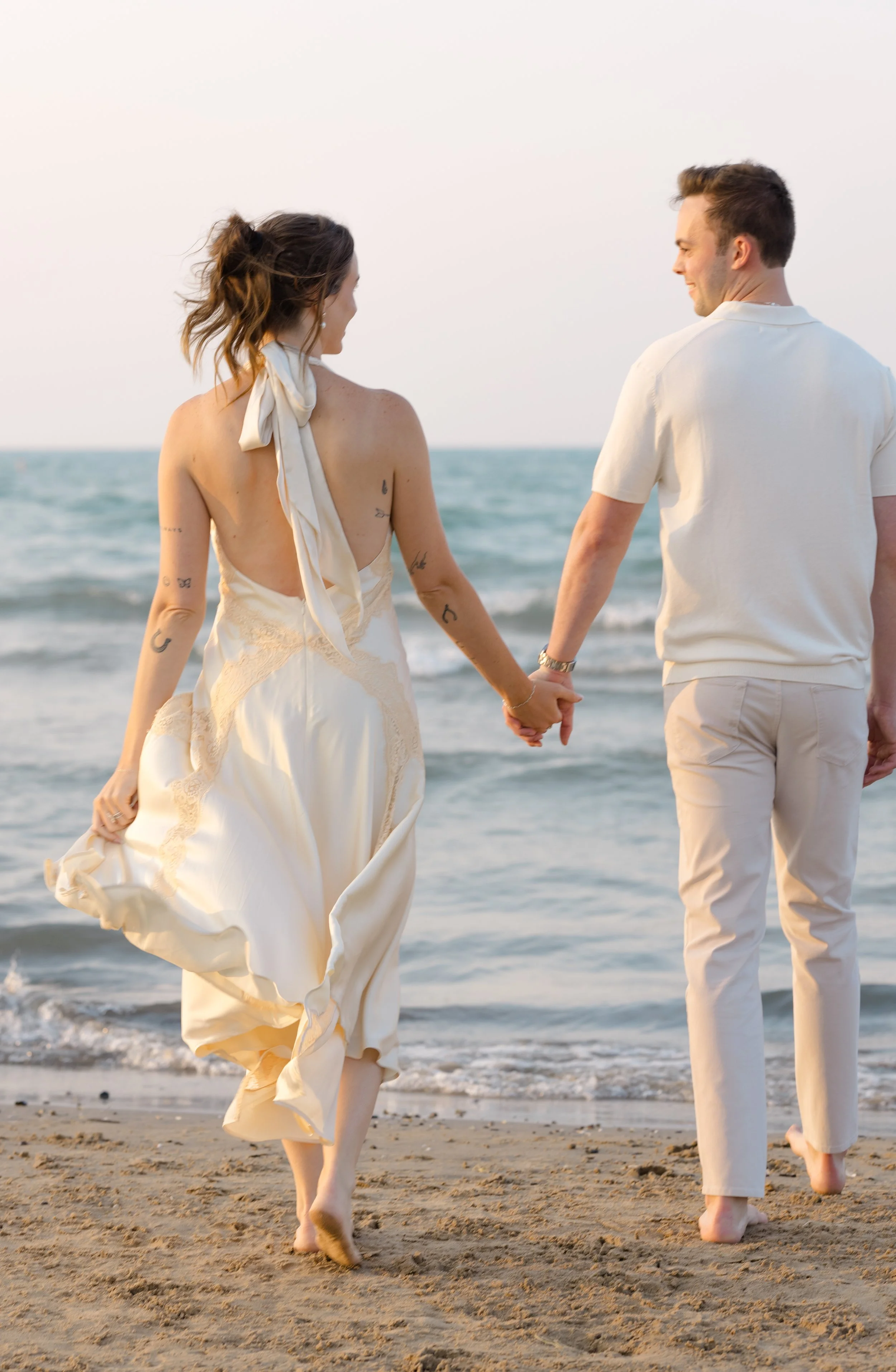 A couple strolling hand-in-hand on the beach during sunset, dressed in light-colored clothing, with their backs to the camera.