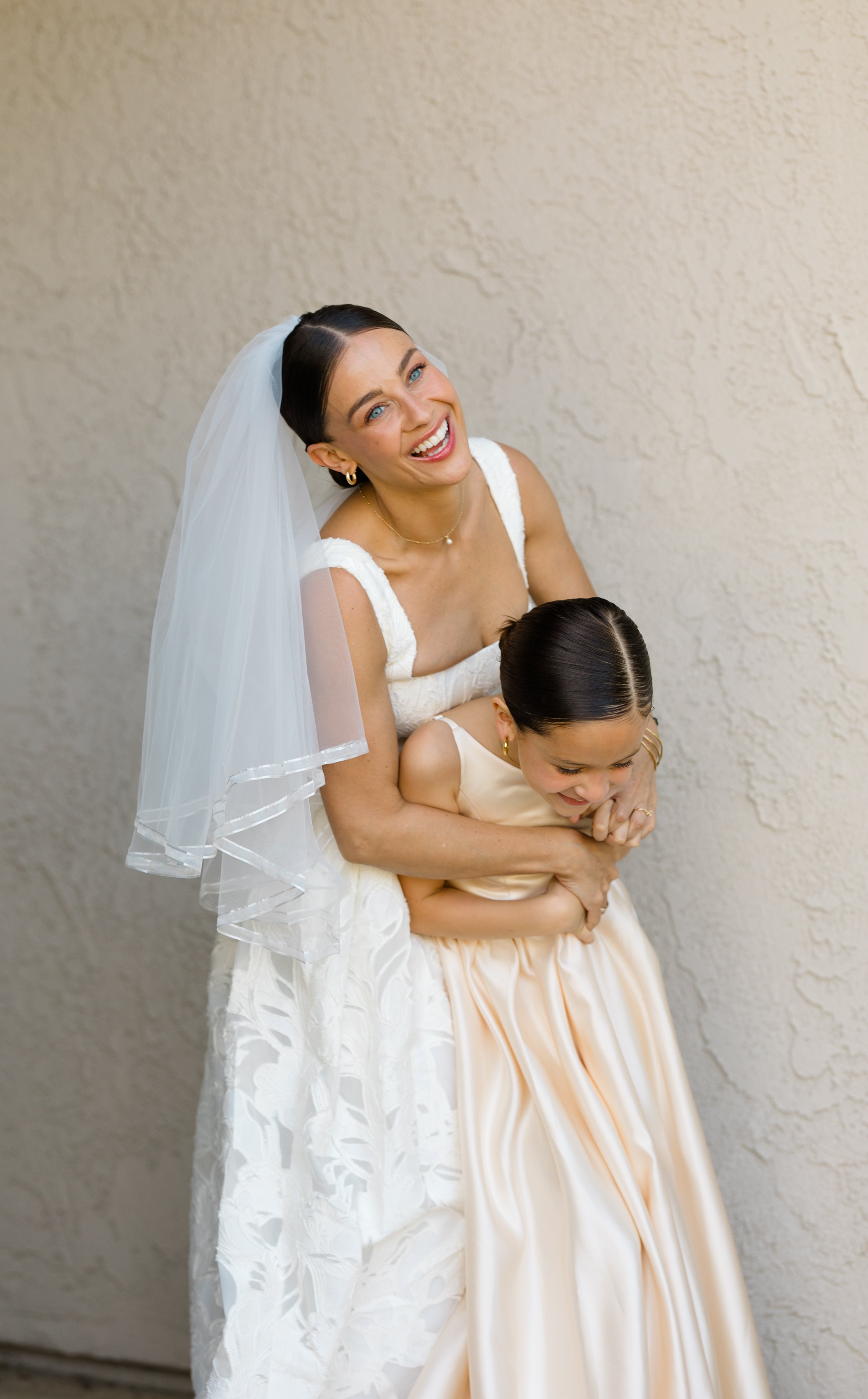 A bride in a white wedding dress and veil hugging a young girl in a cream-colored dress, both smiling happily, standing against a beige textured wall.