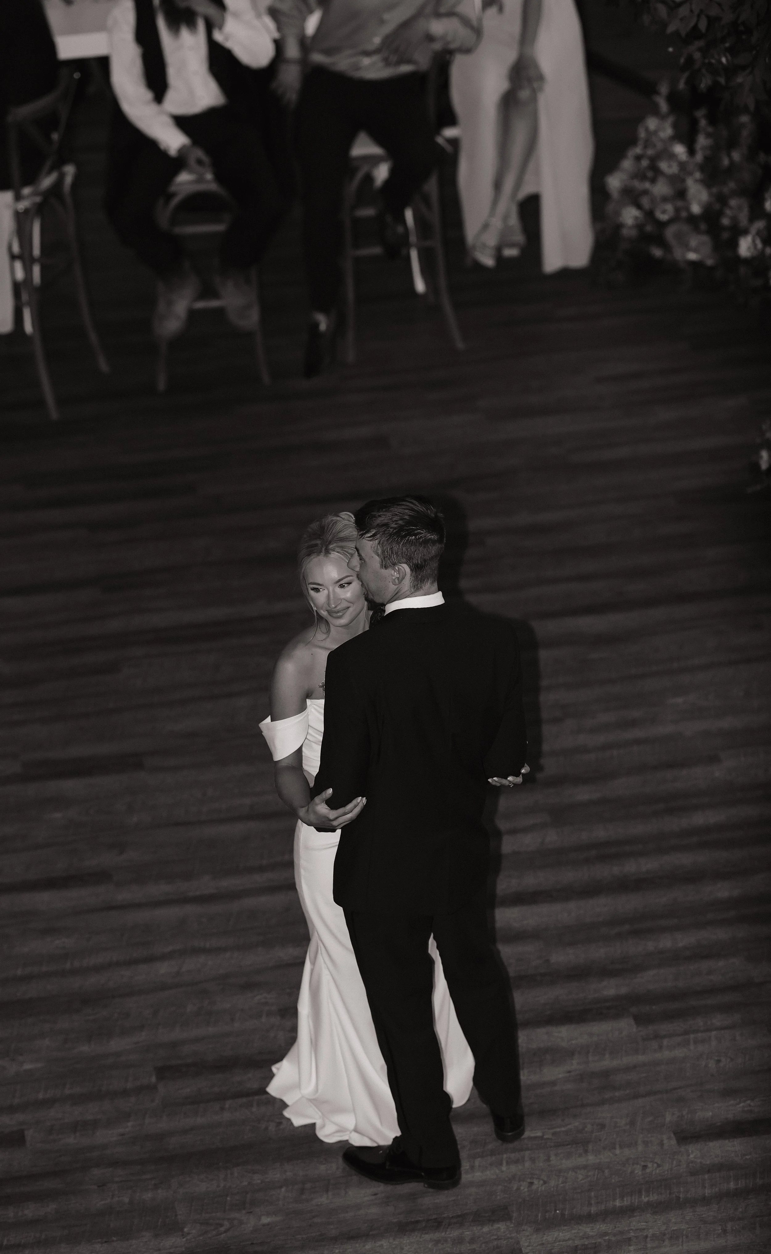 A bride and groom are dancing at their wedding reception on a wooden floor. The bride is wearing a white gown, and the groom is dressed in a black suit. They are close together, holding each other, and looking at each other lovingly. A group of peopl