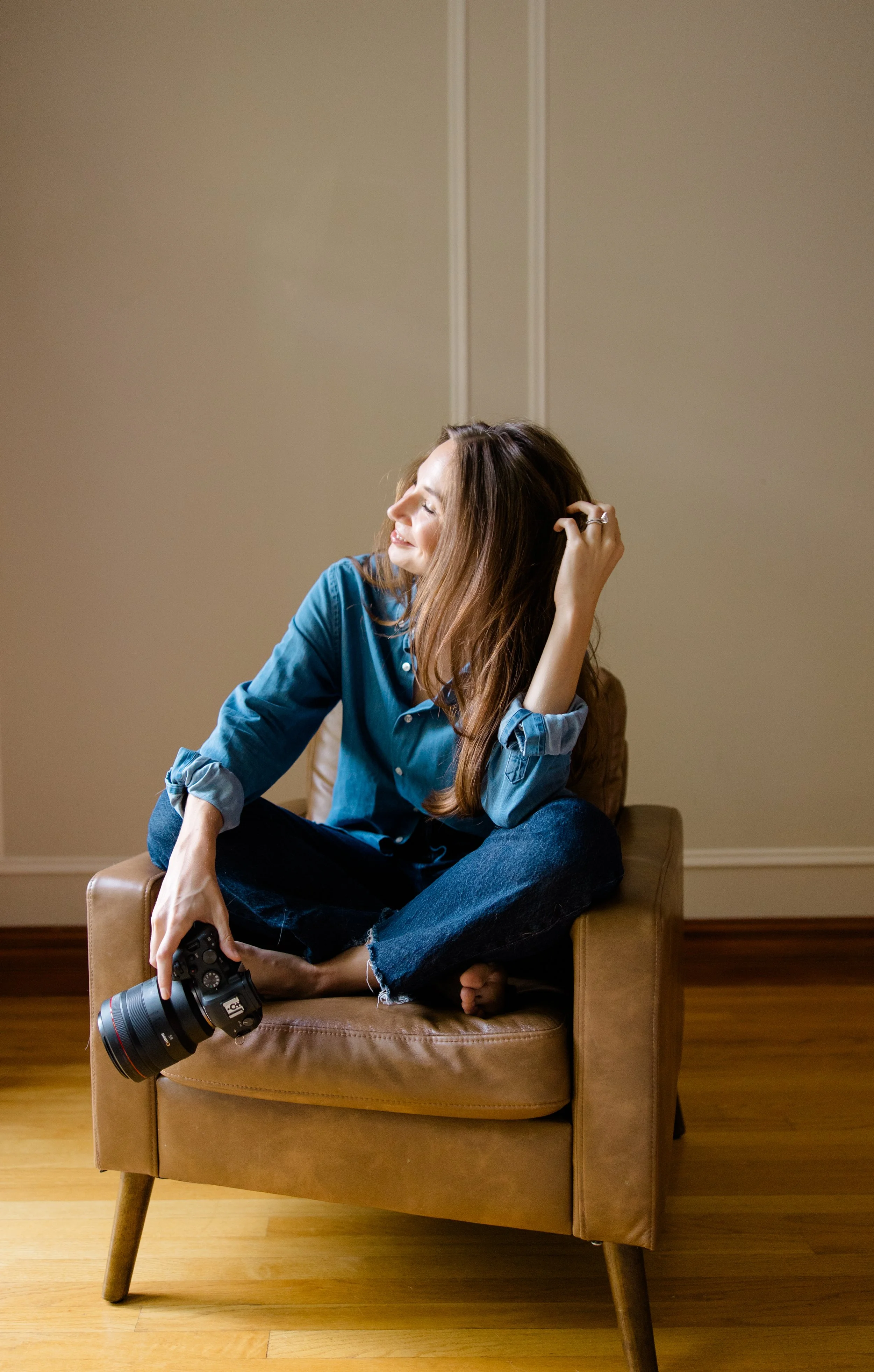 A woman with long brown hair sitting cross-legged on a leather chair, holding a professional camera with a large lens in one hand, and touching her hair with the other, smiling with her eyes closed, in a room with beige walls and wooden floors.