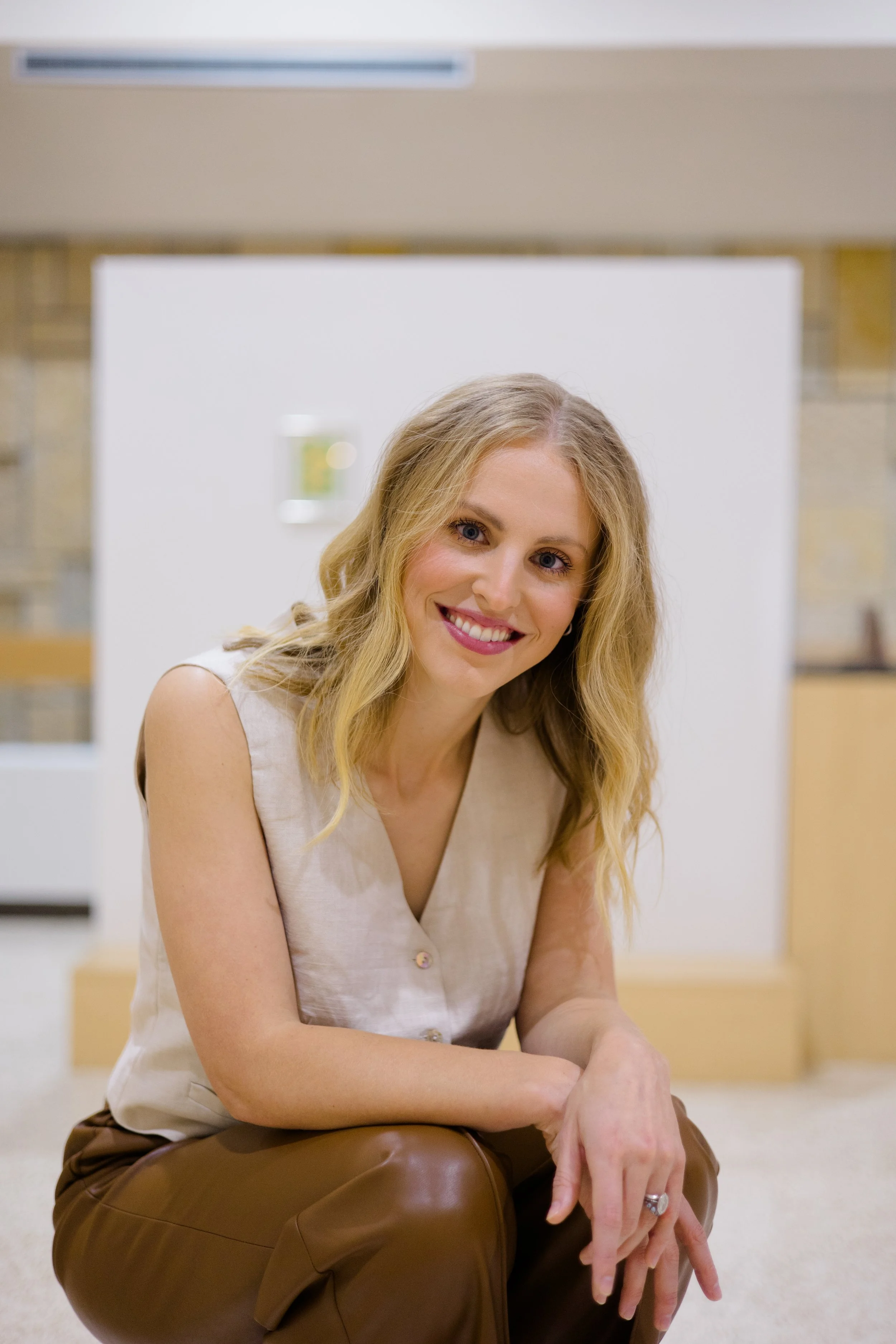 A smiling woman with long blonde hair, wearing a light beige sleeveless top and brown leather pants, sitting on a brown leather chair in an indoor setting with a white wall and an air conditioning unit behind her.