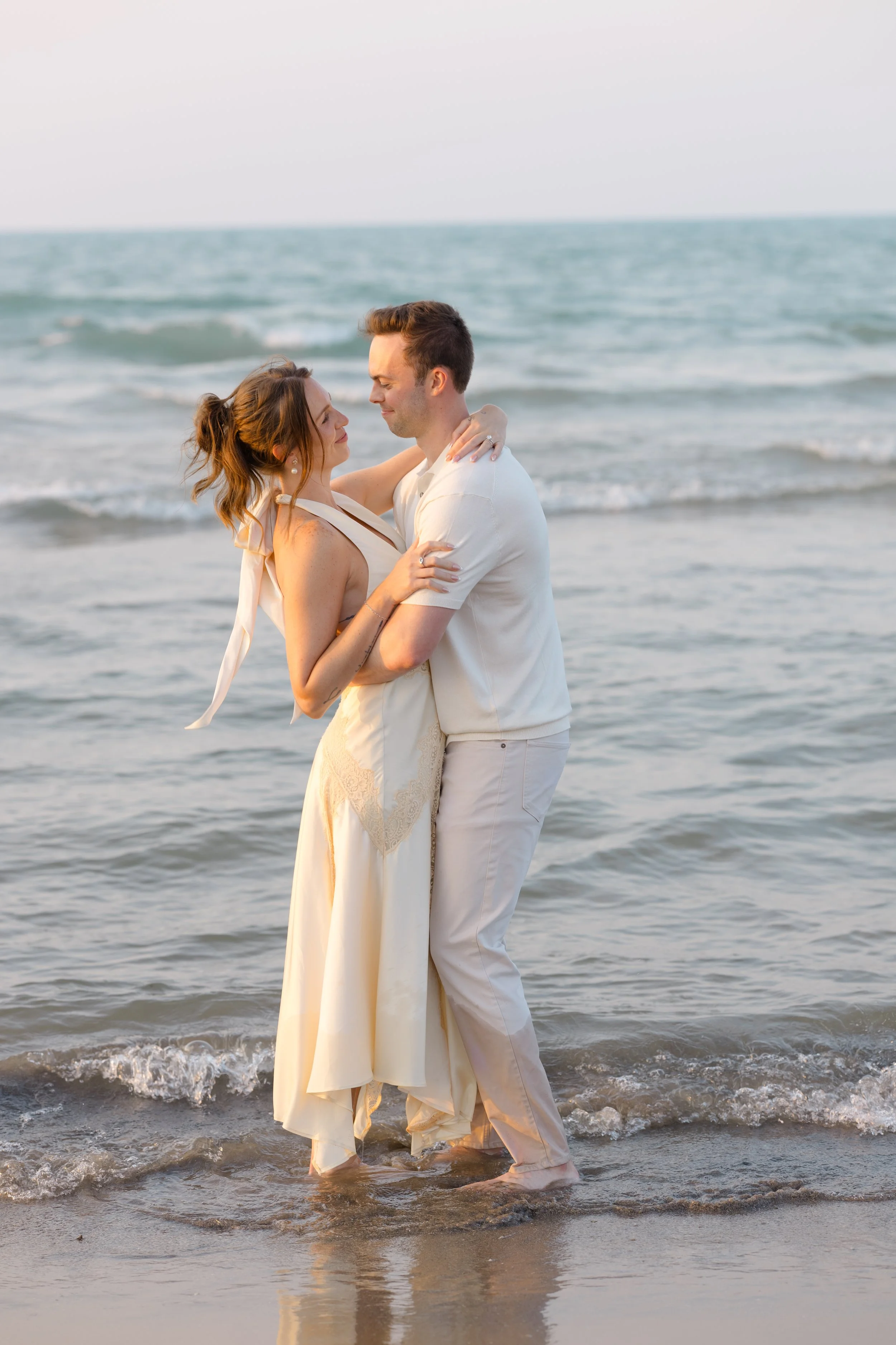 A couple dressed in white, standing in the shallow water of a beach, embracing and looking into each other's eyes during sunset.
