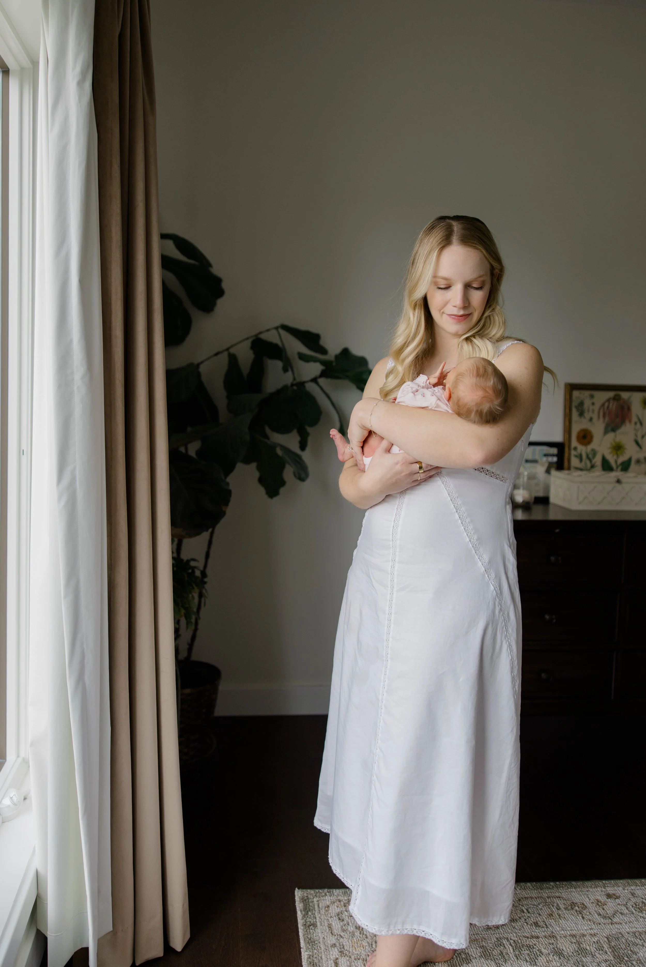 A woman in a white dress holding a baby near a window with curtains and potted plants in a home interior.