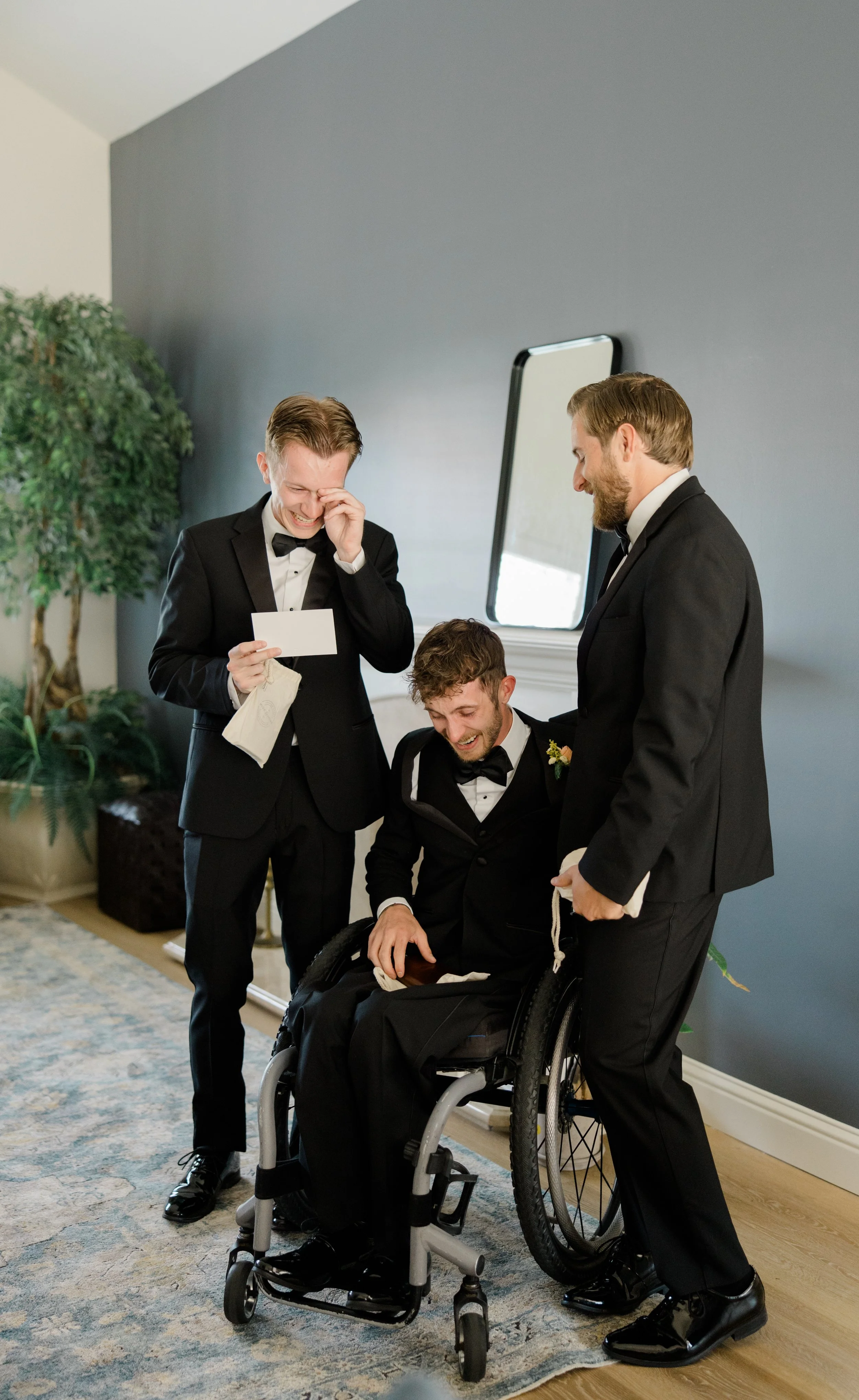 Three men in tuxedos sharing a joyful moment indoors. One man is seated in a wheelchair, while two stand beside him, laughing and smiling. The room has a gray wall, a mirror, greenery, and a patterned rug.