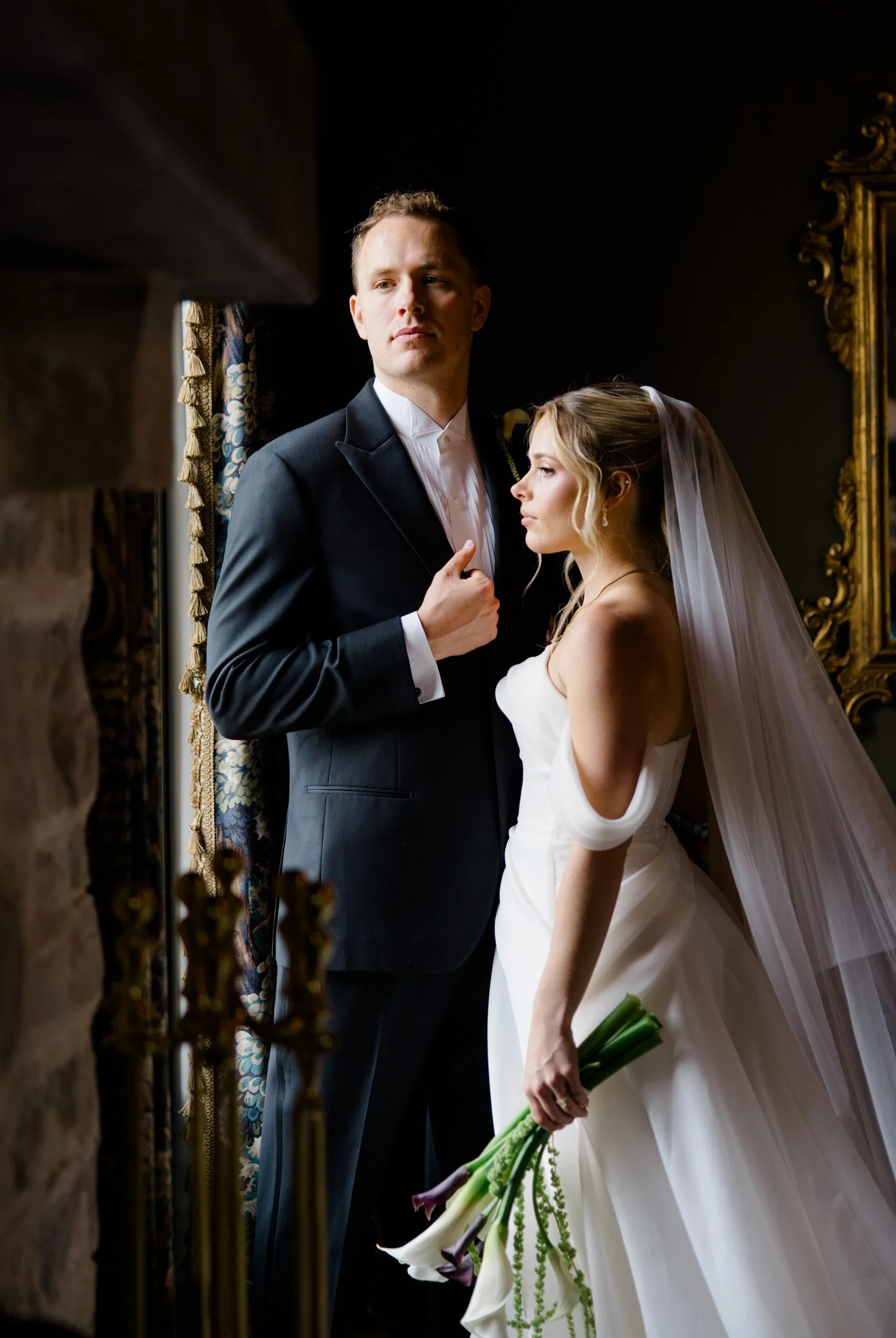 A bride and groom standing indoors, with the bride holding a bouquet of white calla lilies and green flowers. The groom is in a black tuxedo with a white shirt, and the bride is wearing a strapless white wedding dress with a veil. They are near a window with dark curtains a gold-framed mirror on the wall.