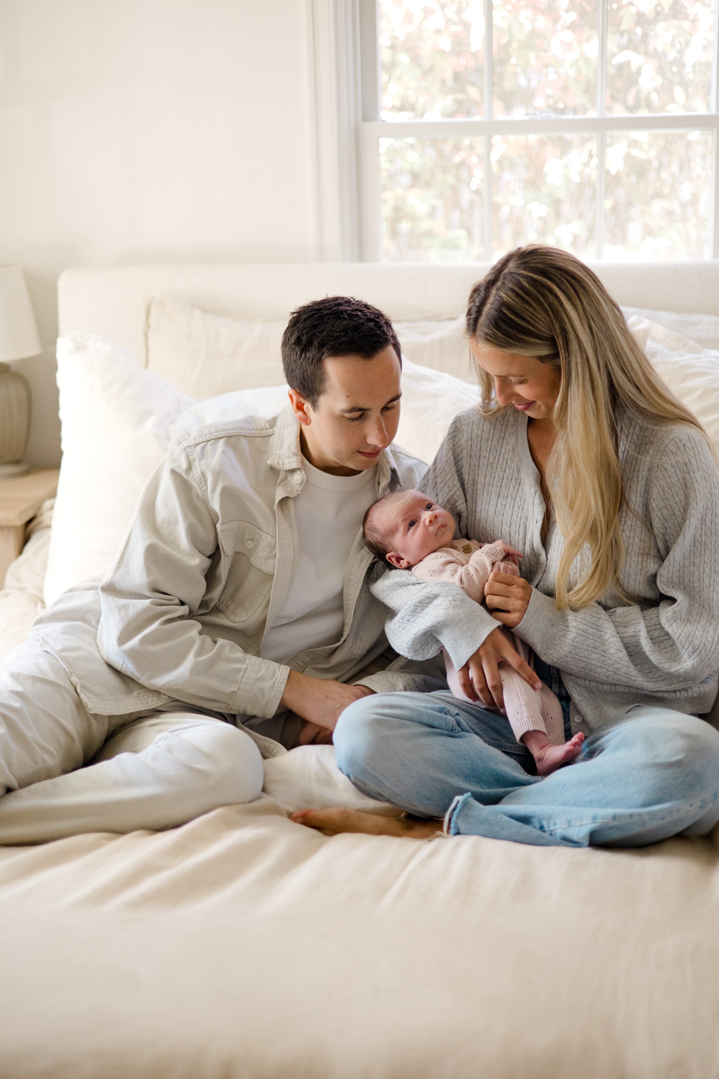 A woman and a man sitting on a bed, holding a newborn baby, in a bright room with large windows and white walls.