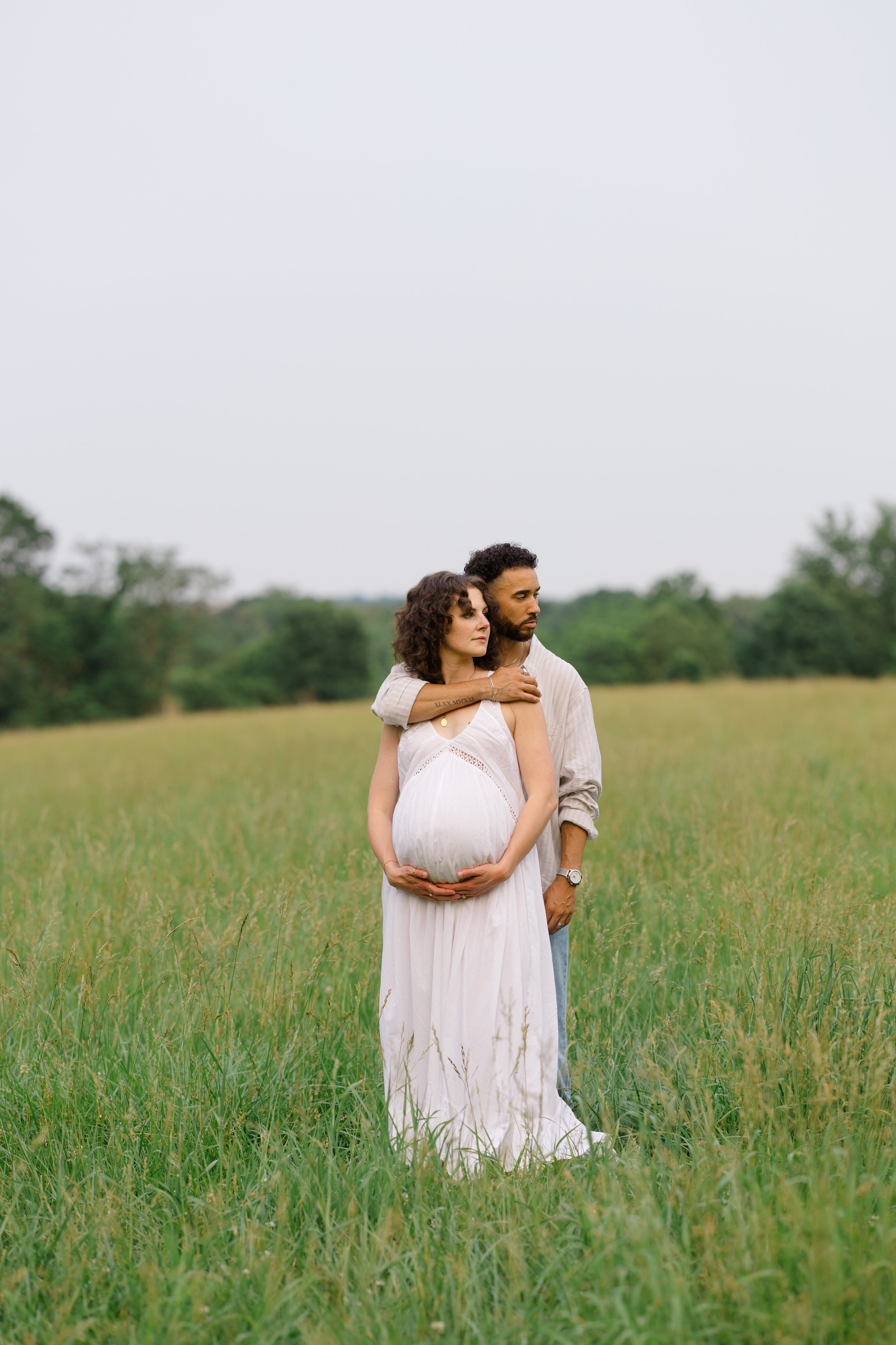 A couple standing in a grassy field, with the woman having a pregnant belly, and the man standing behind her with his arms around her shoulders, both looking to the side.
