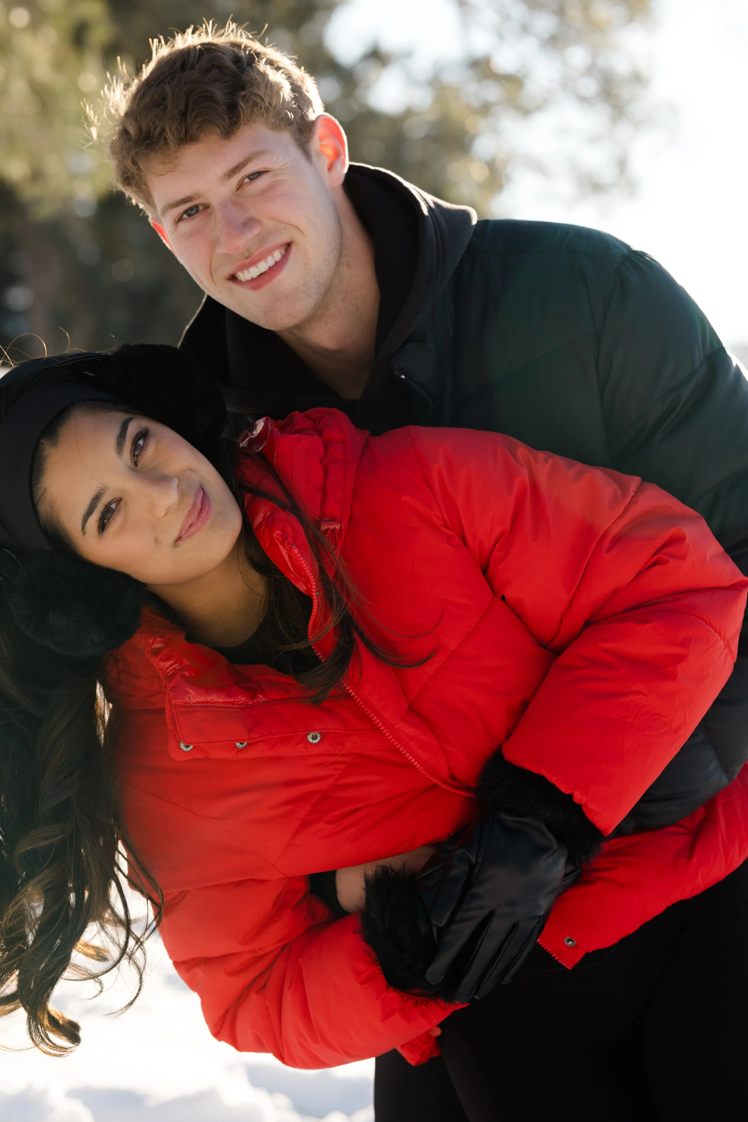 A young man with curly hair and a young woman with dark hair lying in snow, smiling at the camera. The man is wearing a black jacket and the woman a red jacket, both dressed warmly for winter.