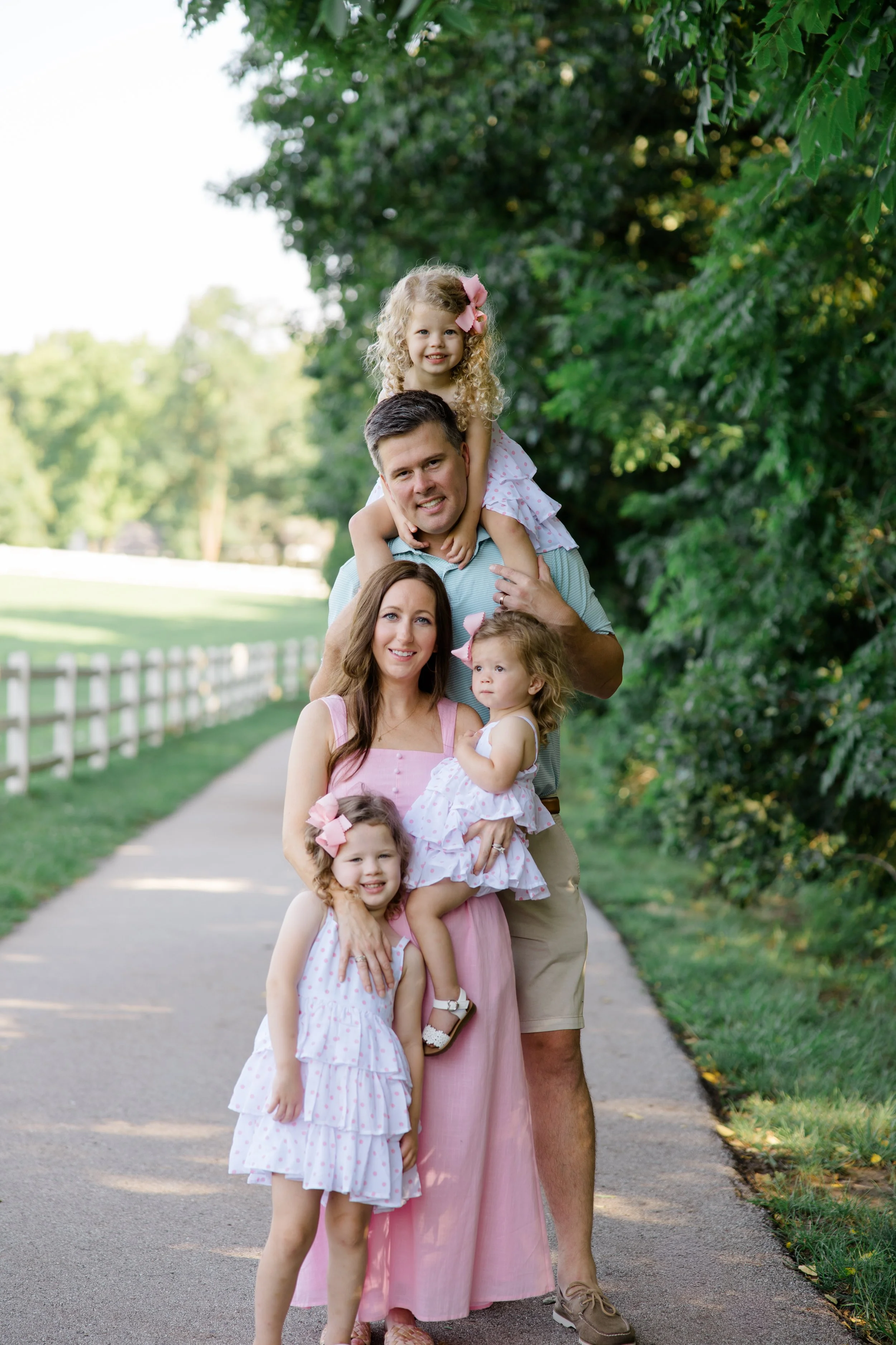A family of six standing on a sidewalk in a park with green trees and a white fence in the background. The family includes a man, a woman, and four young girls. The man is carrying one girl on his shoulders, and the woman is holding another girl in f
