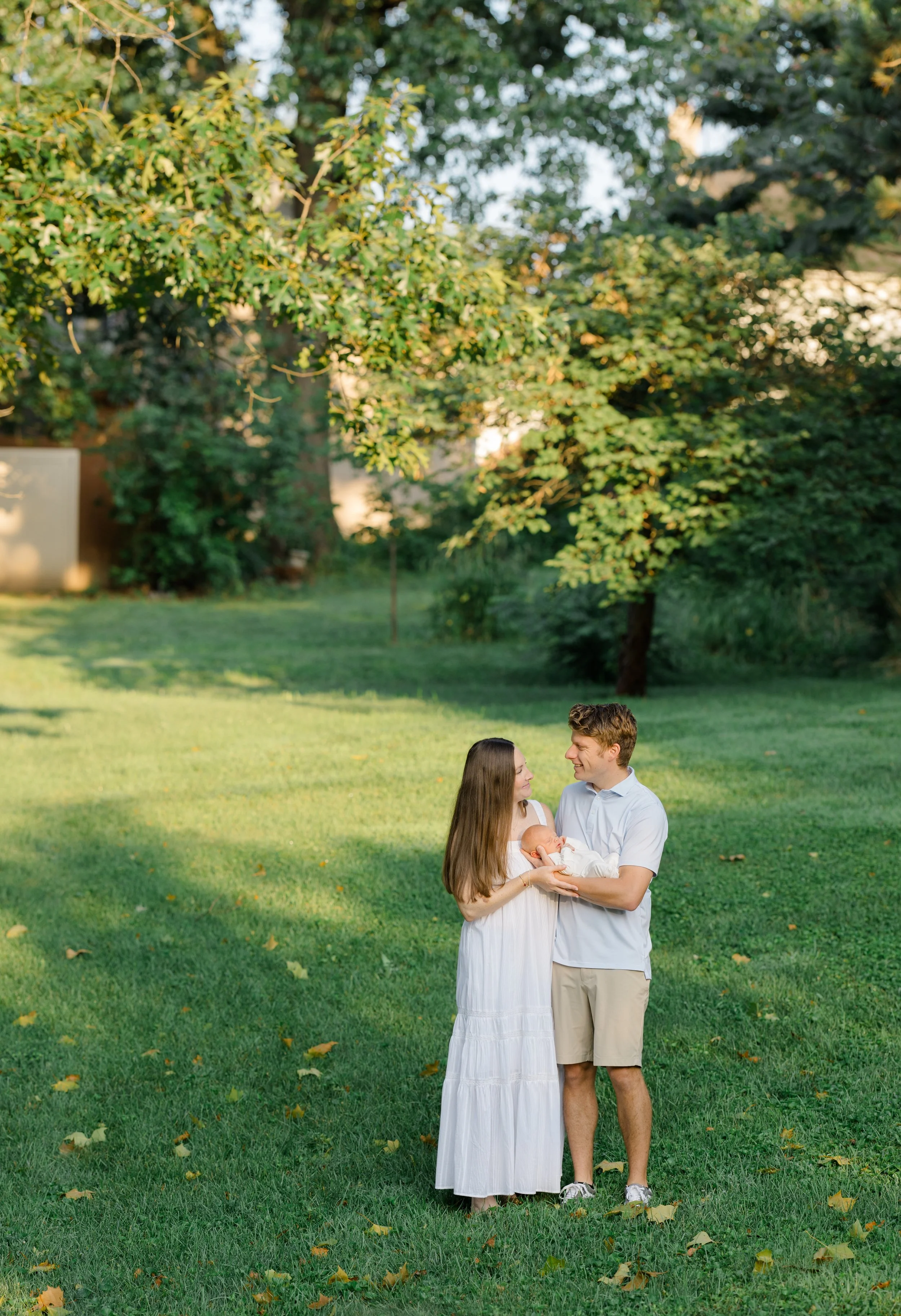 A young couple standing on a green lawn in a park, holding a newborn baby and gazing at each other with trees in the background during daylight.
