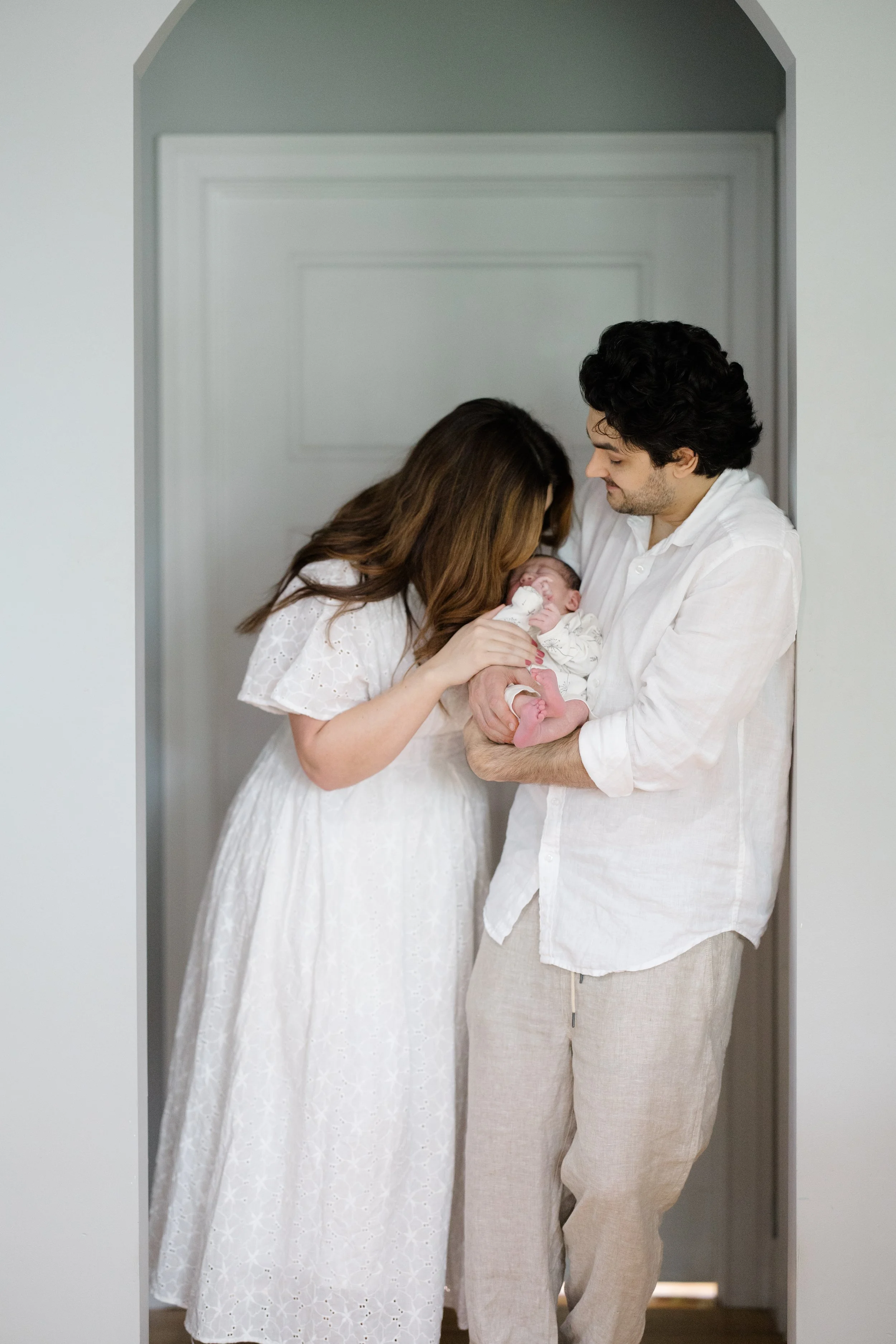 A family of three, a woman, a man, and a newborn baby, standing in a doorway, sharing a tender moment. The woman is wearing a white dress, and the man is dressed in light-colored linen. The woman is gently holding the baby, who is swaddled in white c