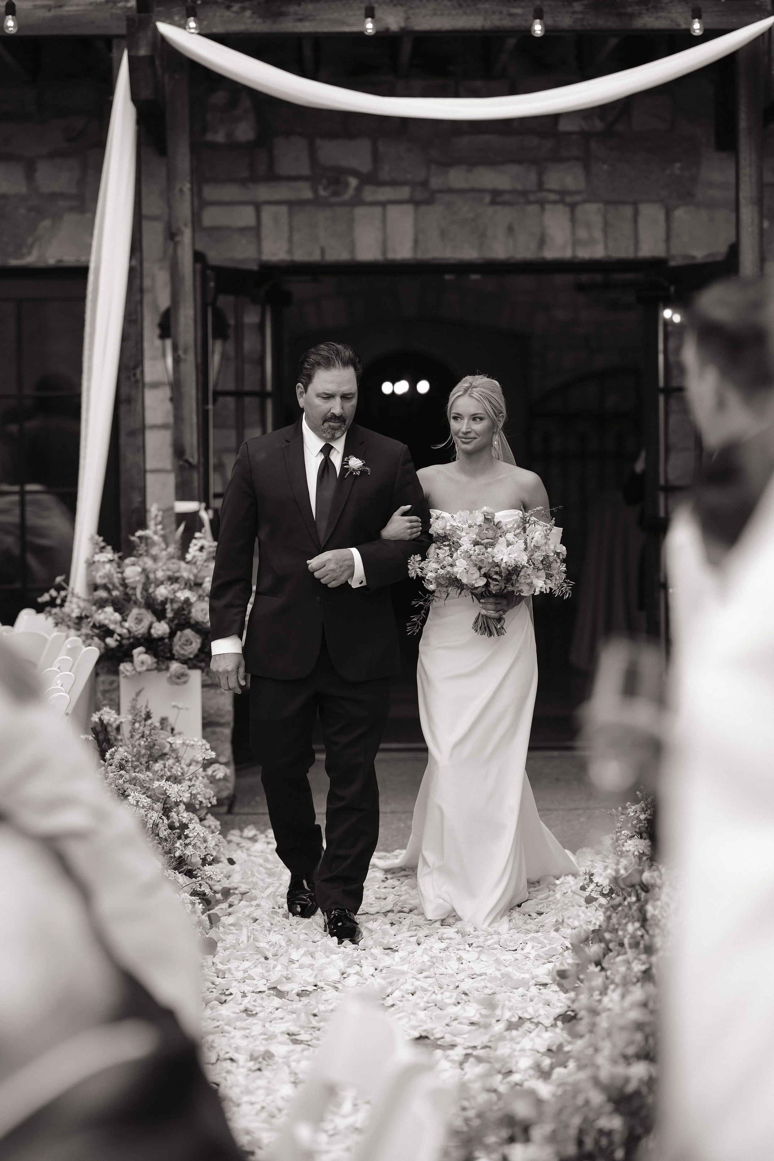 A bride in a strapless white wedding dress holding a bouquet of flowers, walking down the aisle with a man in a black suit, presumed to be her father, in a rustic reception hall decorated with flowers and white drapes.