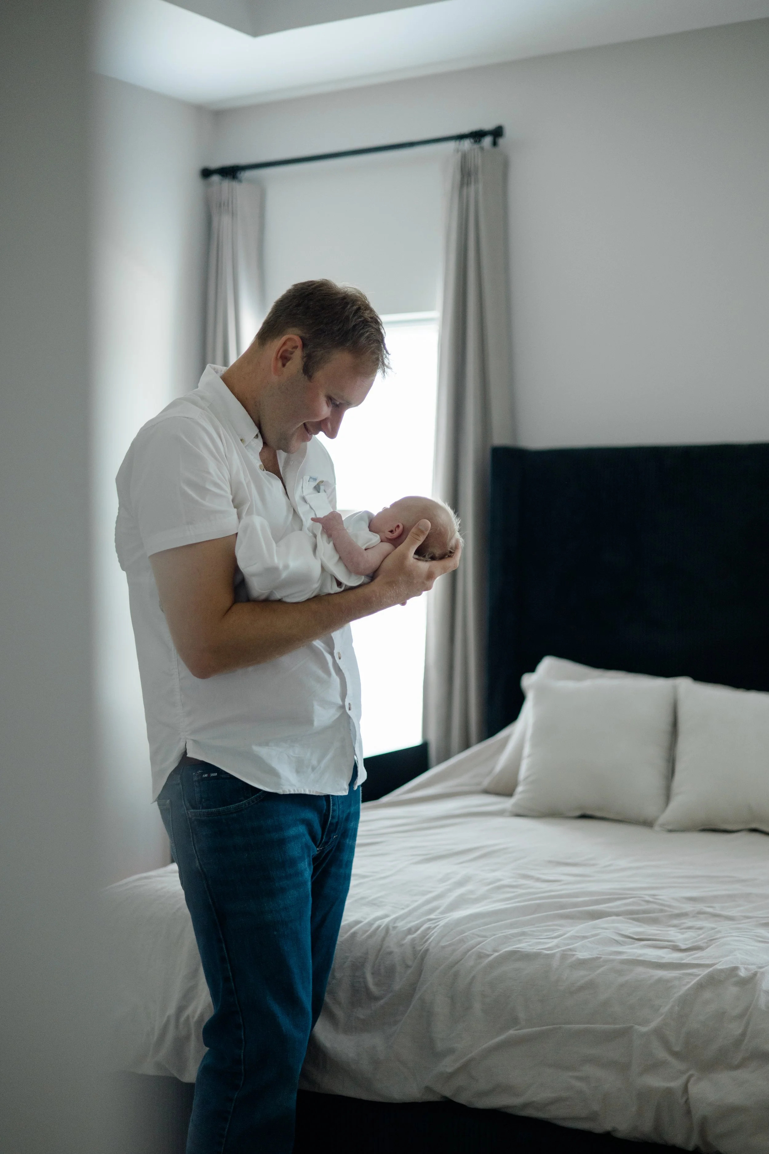 A man holding a baby in a bedroom with a bed, pillows, and curtains, illuminated by natural light from a window.