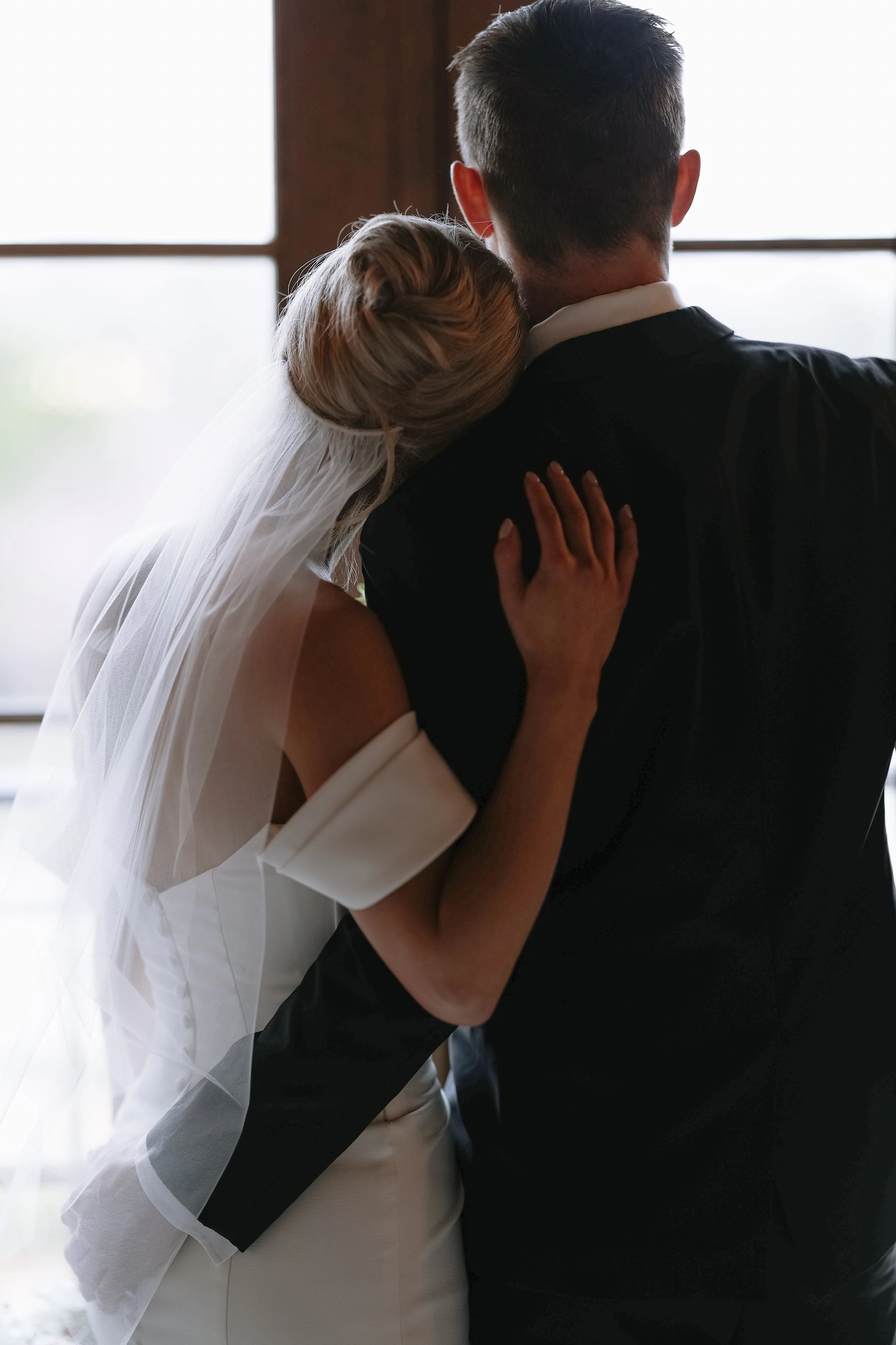 A bride and groom embrace each other, seen from the back, standing near a window with bright light outside. The bride is wearing a white dress and veil, and the groom is in a dark suit with short, styled hair.