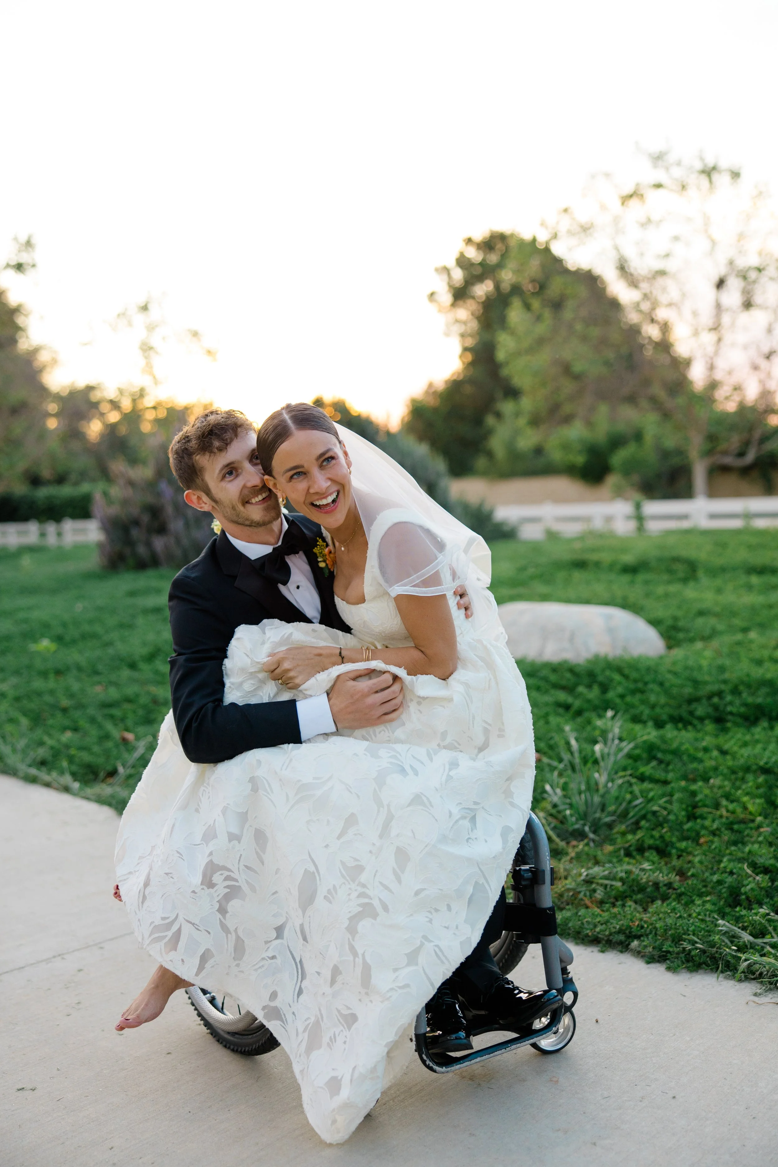 A bride and groom smiling outdoors during sunset, with the bride in a wheelchair, in a park with trees and a white fence in the background.