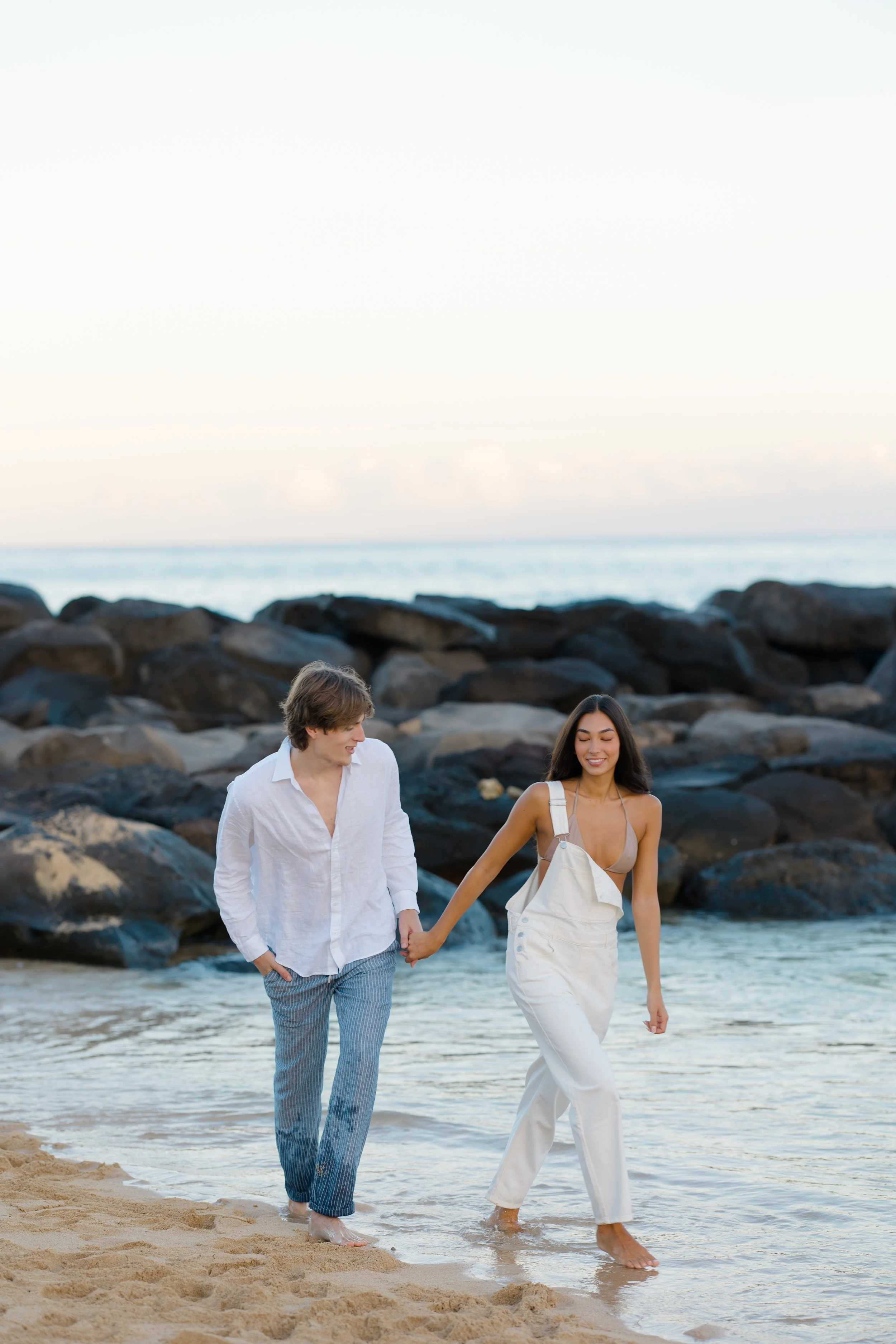 A couple walking hand in hand along the beach shore, with ocean and rocks in the background during sunset.