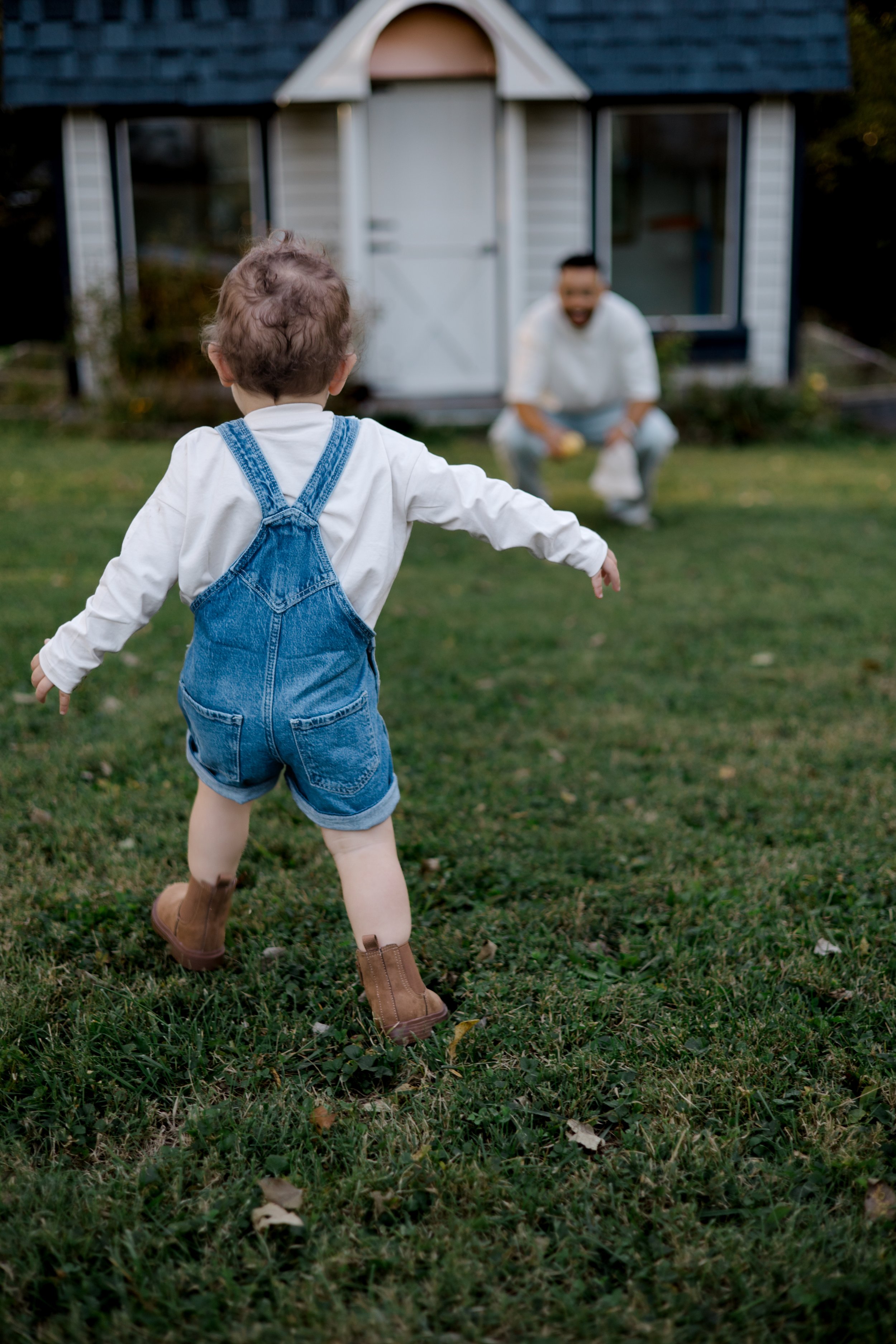 A young child in denim overalls and brown boots running towards an adult who is crouching near a small shed in a backyard, with a grassy area and trees in the background.