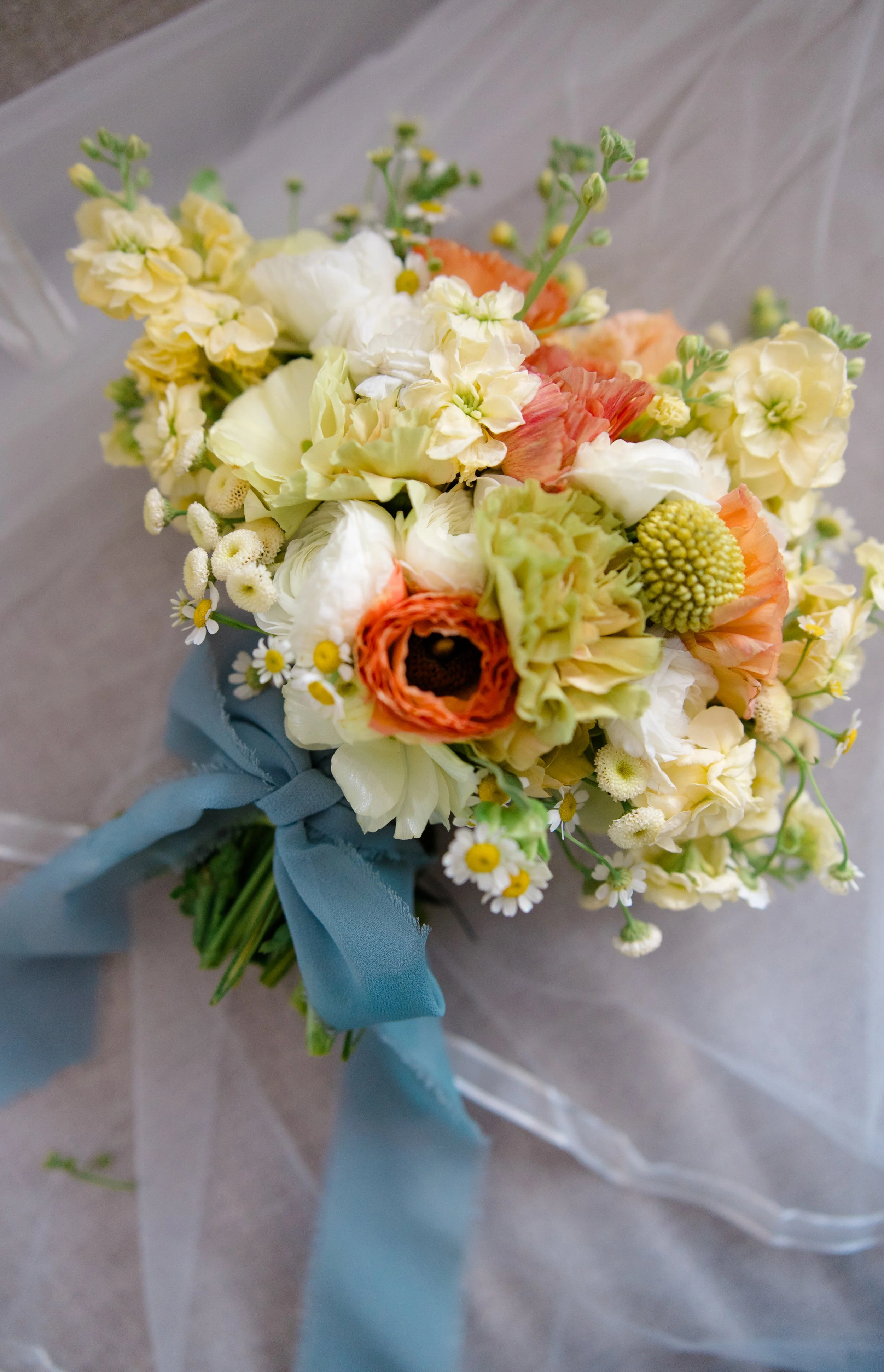 A bouquet of mixed flowers including white, cream, peach, and green blossoms, tied with a light blue ribbon, resting on a gray surface.
