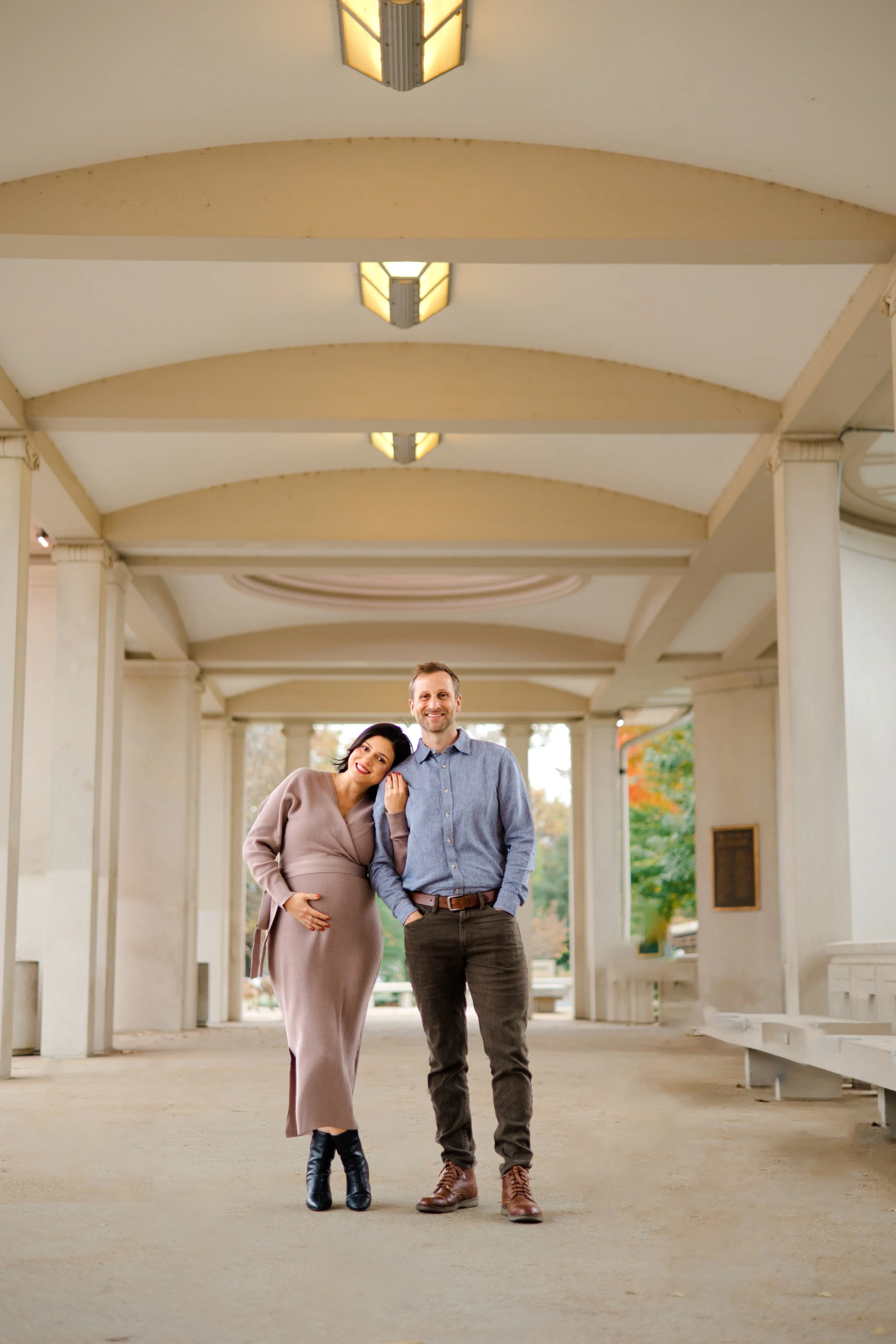 A pregnant woman and a man standing close together in an open-air corridor, smiling. The woman is wearing a beige dress with black boots, and the man is wearing a blue shirt with brown pants and boots.