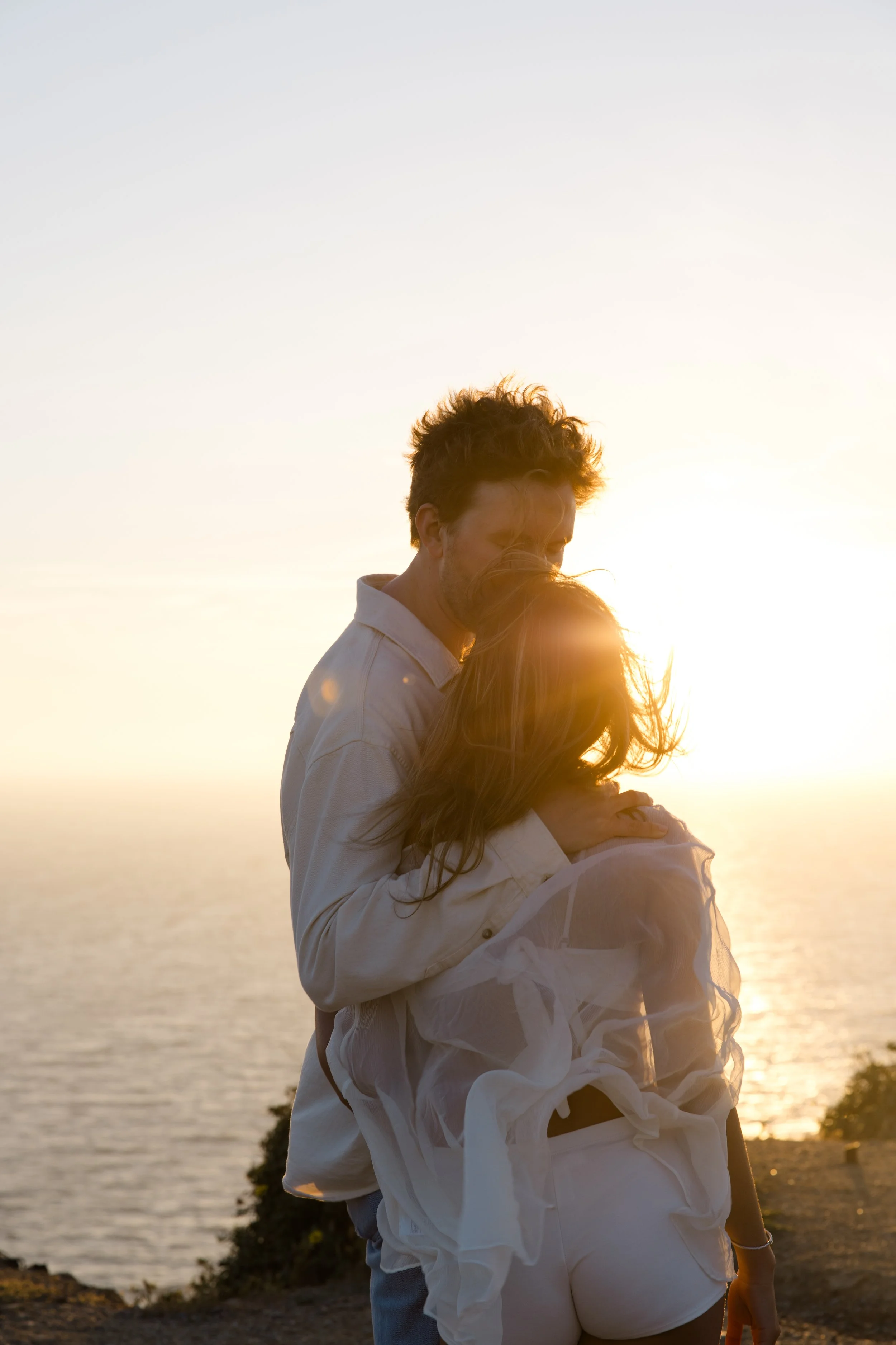 A couple kisses at sunset by a body of water, with the woman's hair blowing in the wind and the sunlight creating a warm glow.