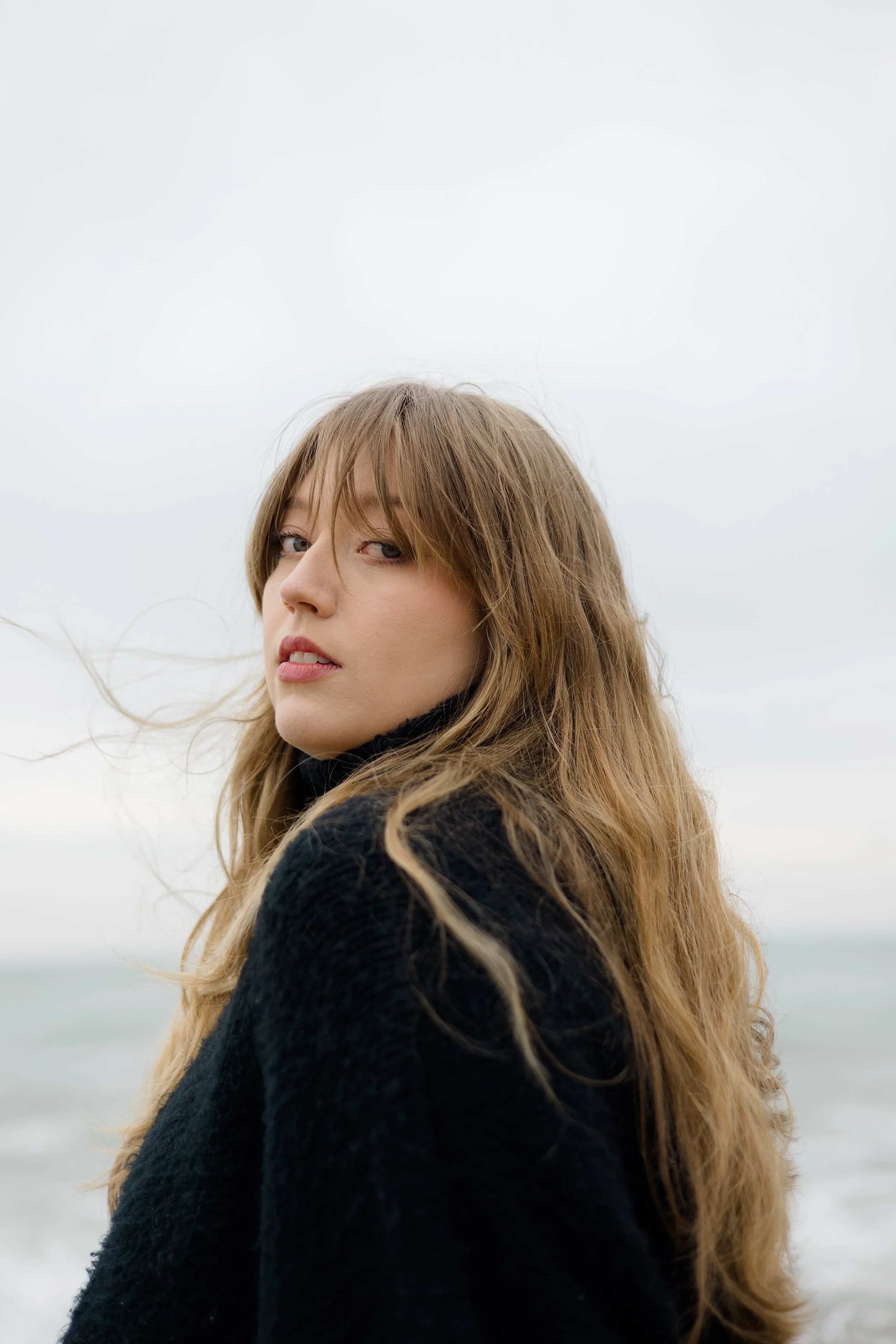 A young woman with long, wavy brown hair looking over her shoulder at a beach with a cloudy sky.
