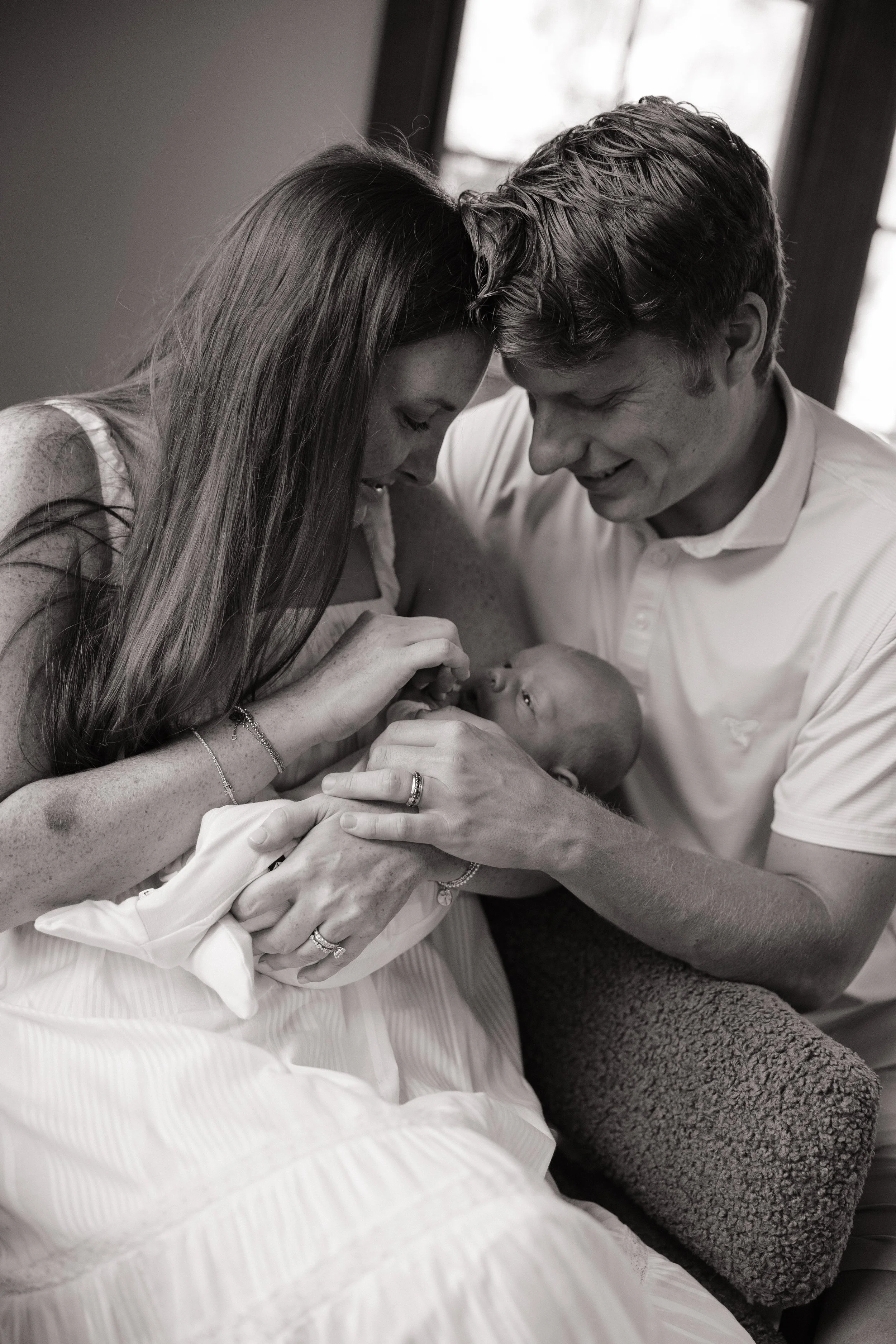A couple, a woman with long hair and a man with short hair, holding and looking at their newborn baby, who is cradled in a woman’s arms. The scene appears to be in a hospital or birthing center, illuminated by natural light from a window.