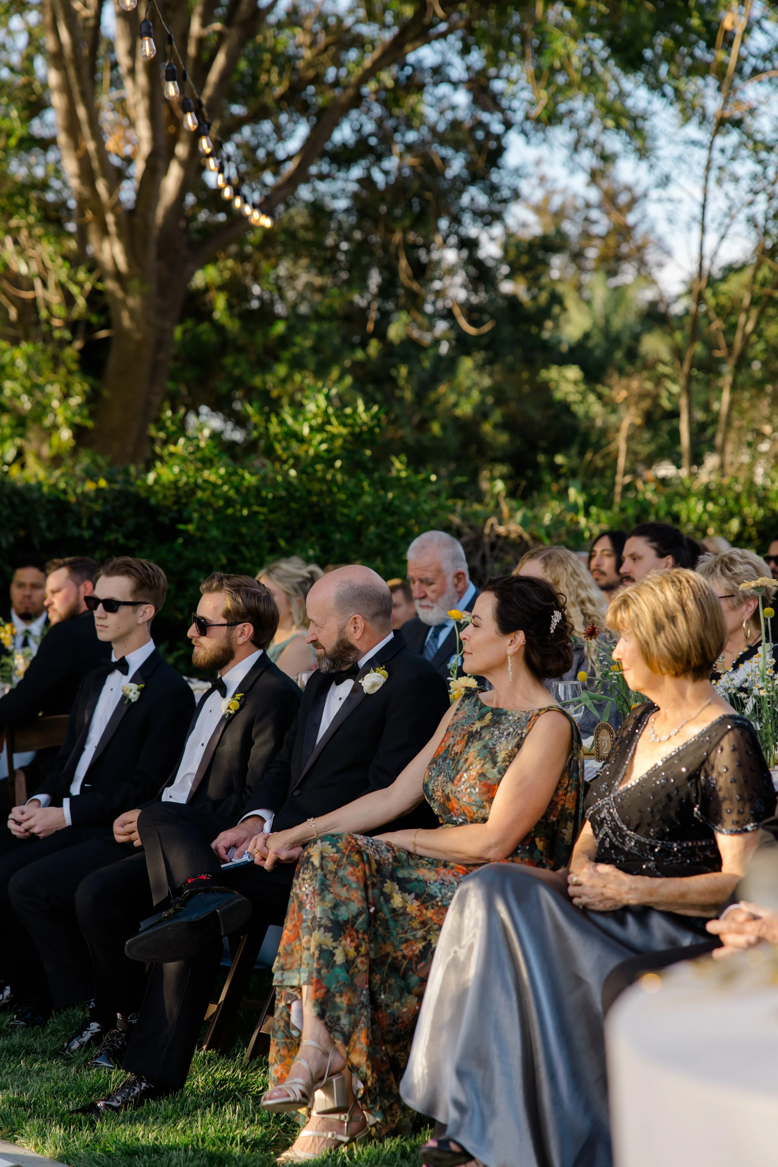 A group of people dressed in formal wear seated outdoors at a wedding or event, with trees and string lights in the background.