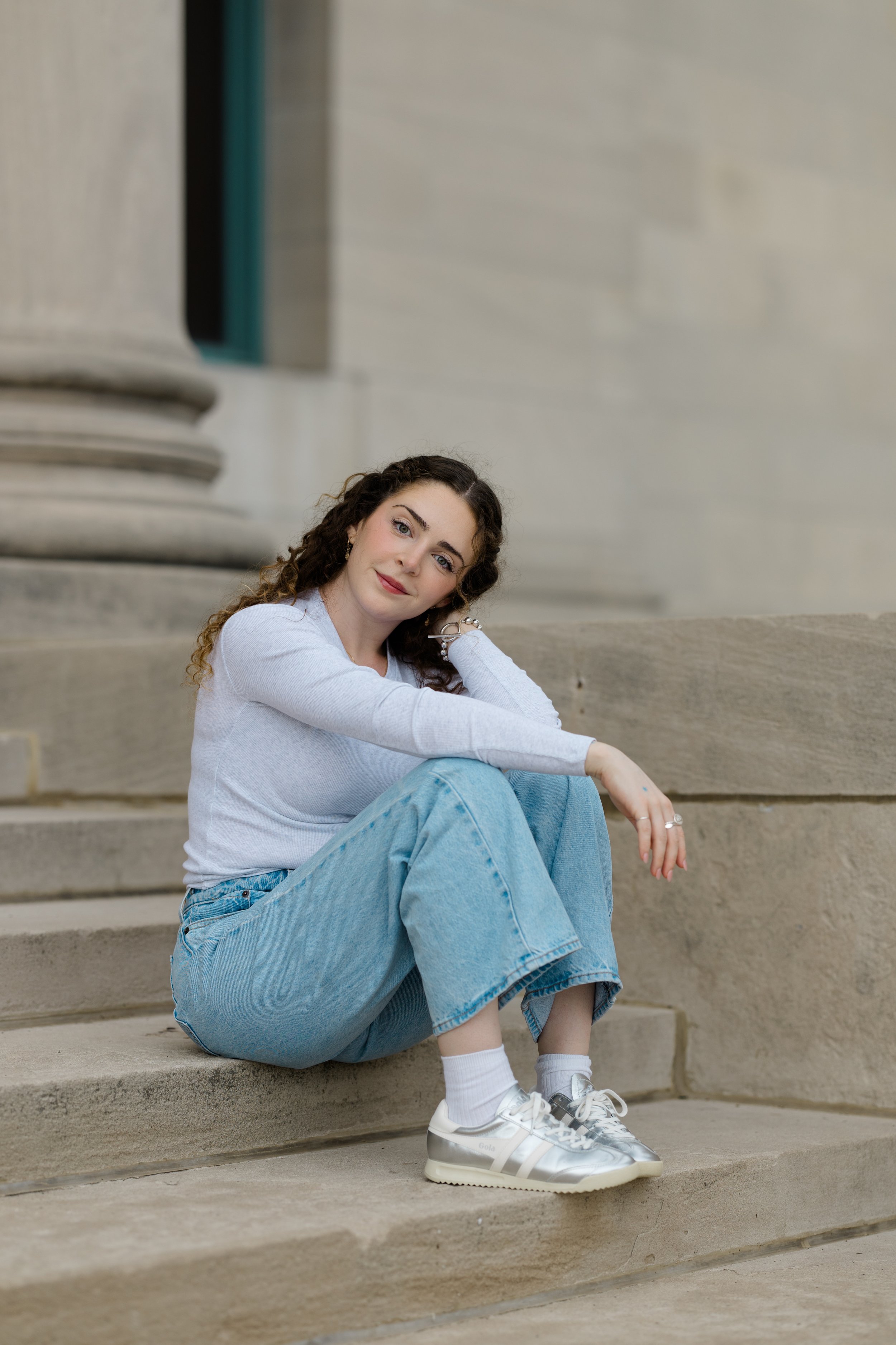 A young woman with curly brown hair sitting on steps outside, wearing a white long-sleeve shirt, blue jeans, and silver sneakers, looking at the camera with a slight smile.