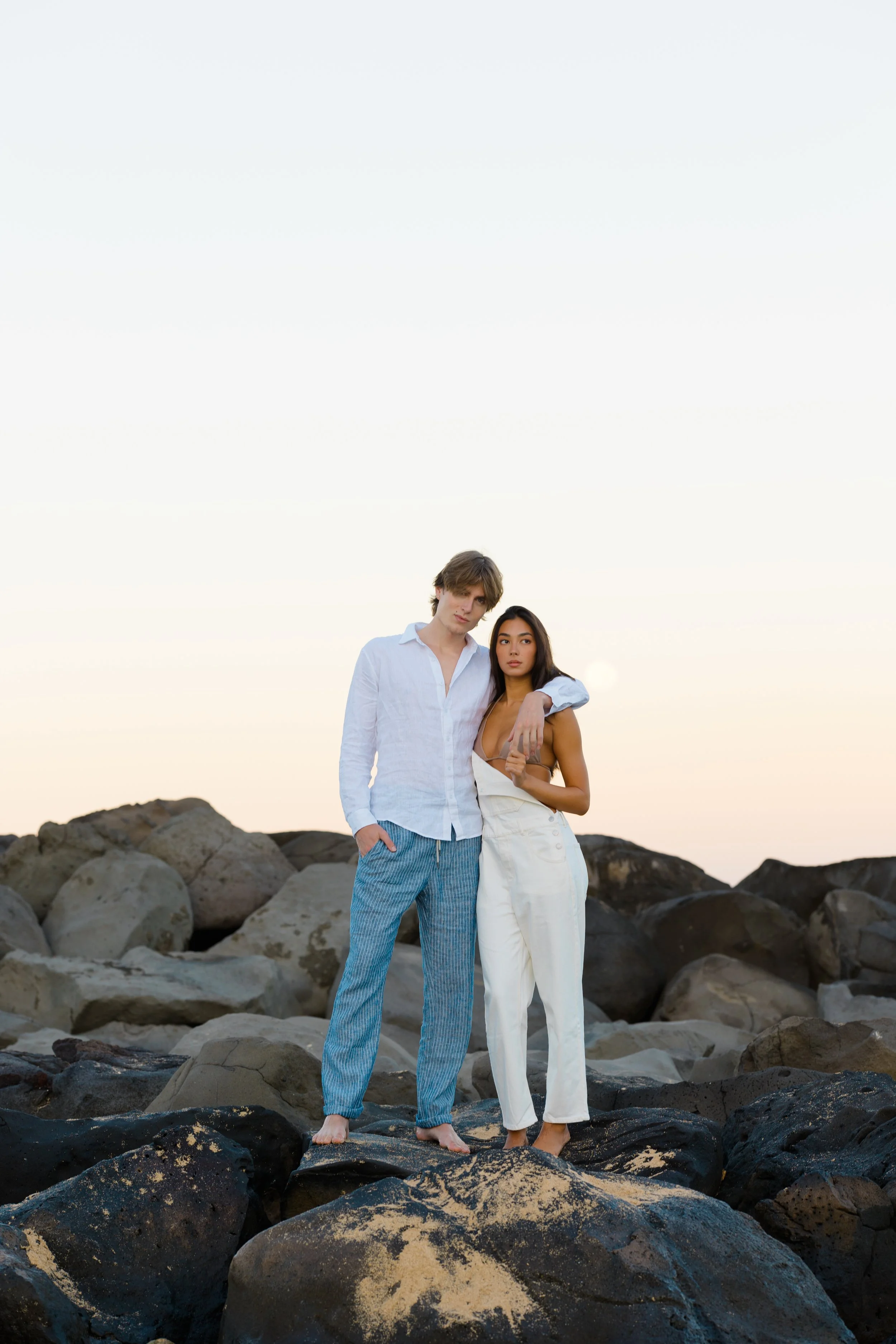 A young man and woman standing close together on a rocky beach with the ocean at their feet during sunset. The man has light brown hair, wears a white shirt and blue striped pajama pants, and has his hand in his pocket. The woman has dark hair, wears