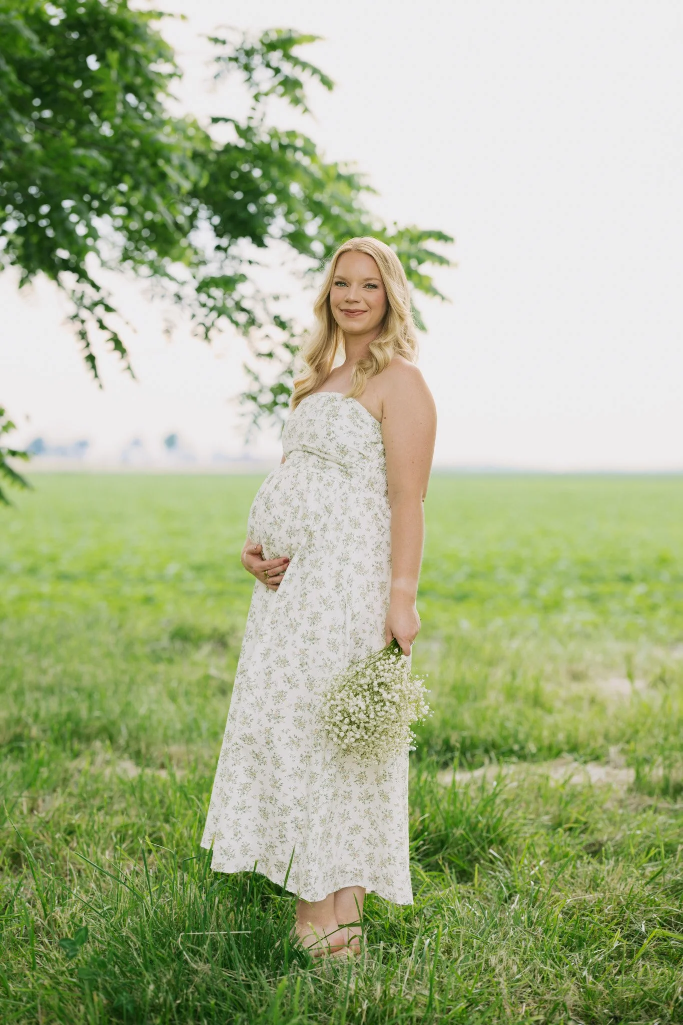 A pregnant woman in a floral dress holding a bouquet of white flowers standing in a grassy field with trees in the background.