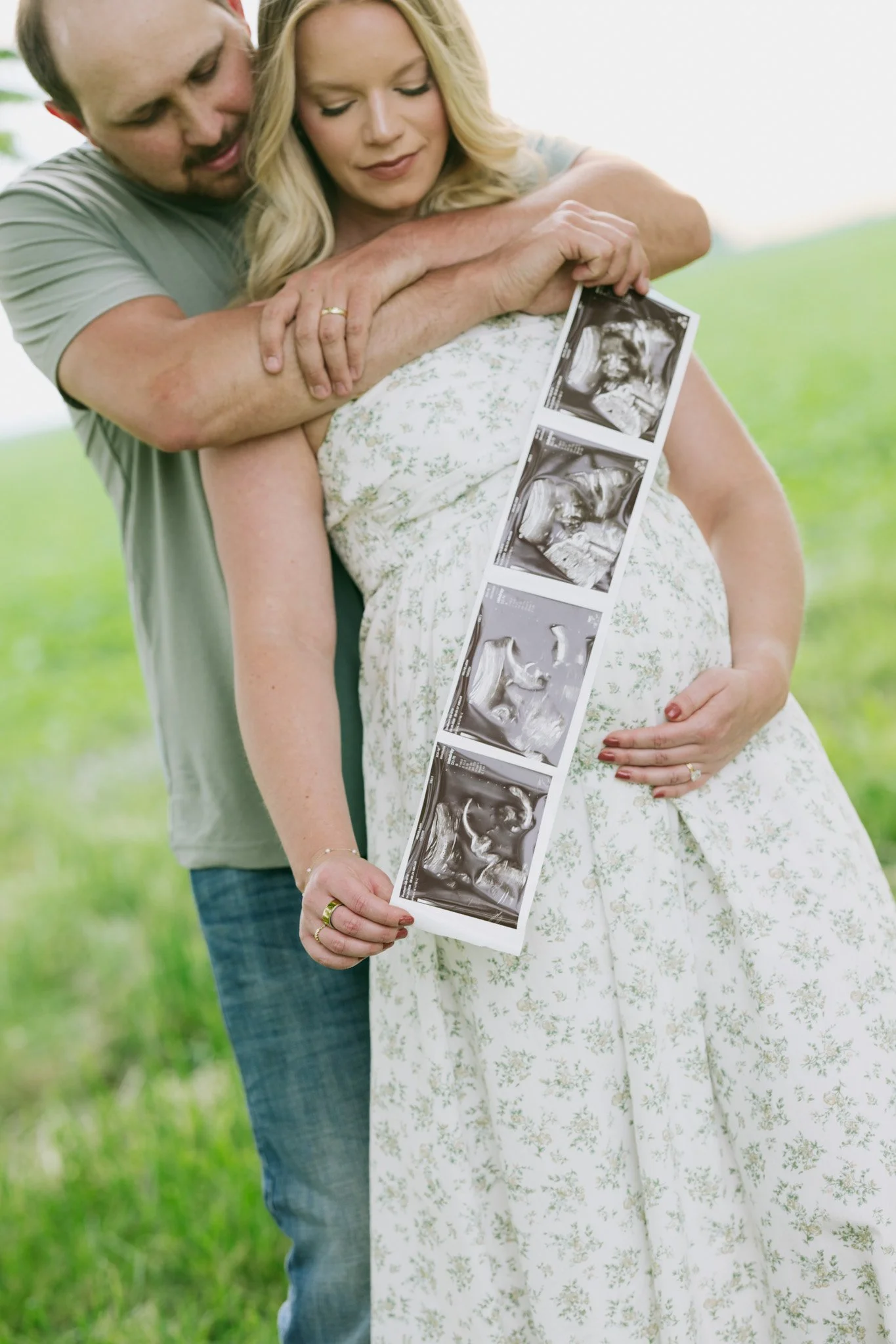 A pregnant woman in a white floral dress holding ultrasound images, with a man embracing her from behind in a grassy outdoor setting.