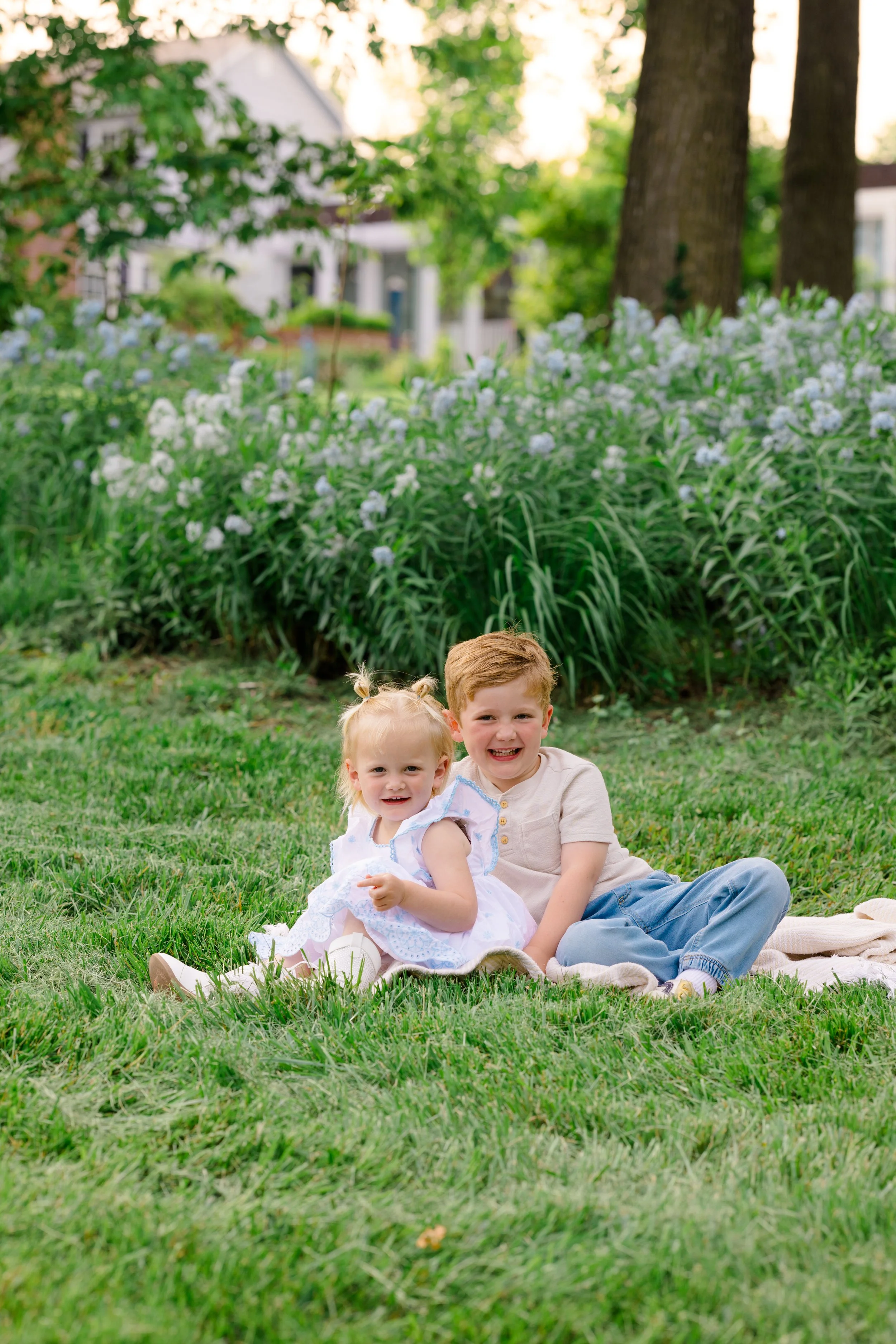 A young boy and girl sitting on a grassy lawn, smiling, with trees and a house in the background.