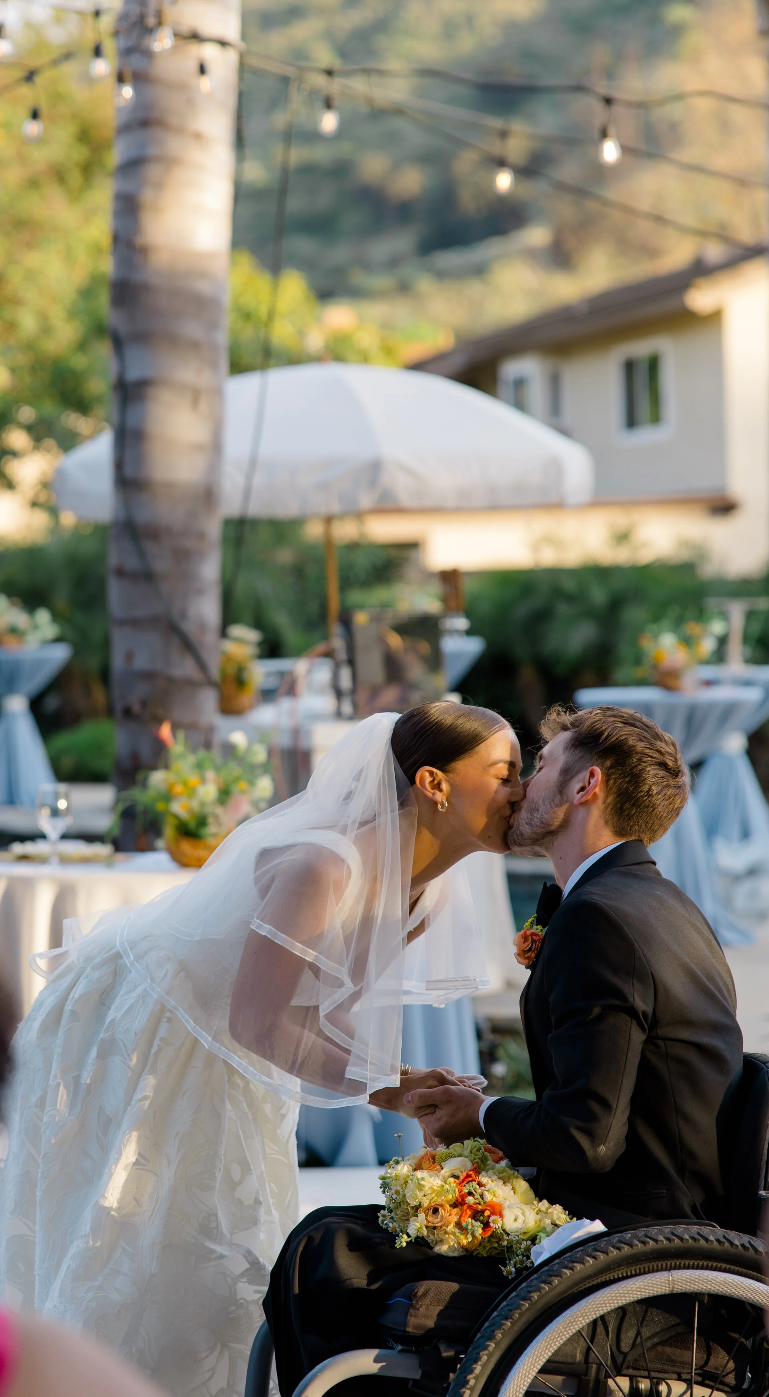 A bride and groom share a kiss at their outdoor wedding ceremony. The bride is wearing a white wedding dress and a veil, while the groom is dressed in a black tuxedo. They are surrounded by tables and decorations in a garden setting, with string ligh