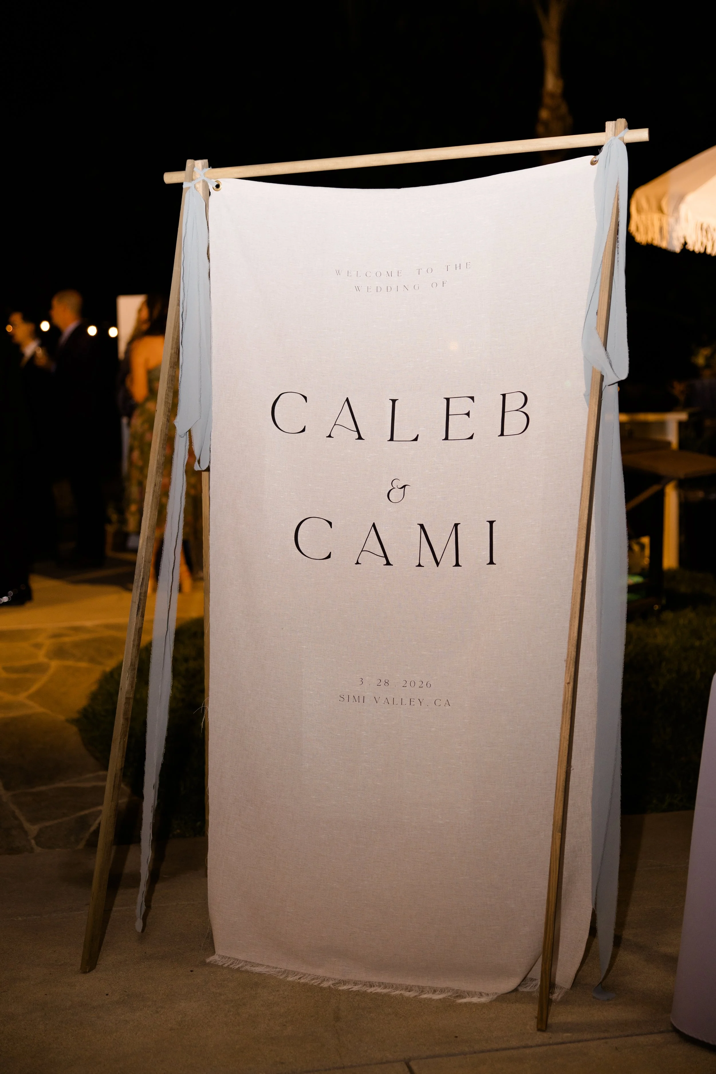A wedding sign with elegant black text reads: 'Welcome to the wedding of Caleb & Cami, 3.28.2026, Simi Valley, CA,' displayed outdoors at night with blurred guests and warm lighting in the background.