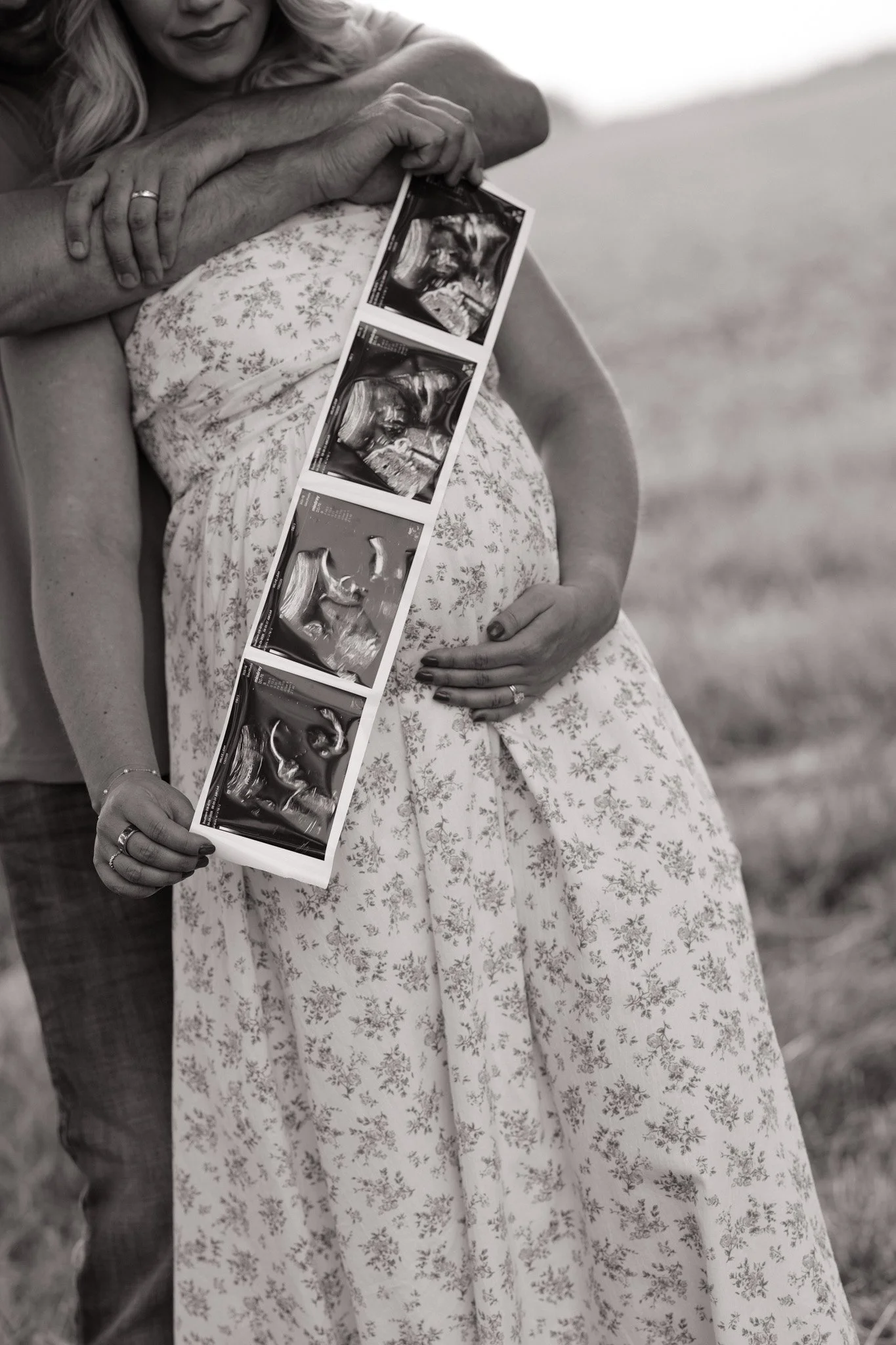 A pregnant woman in a floral dress holding ultrasound images, with a person standing behind her, outdoors.