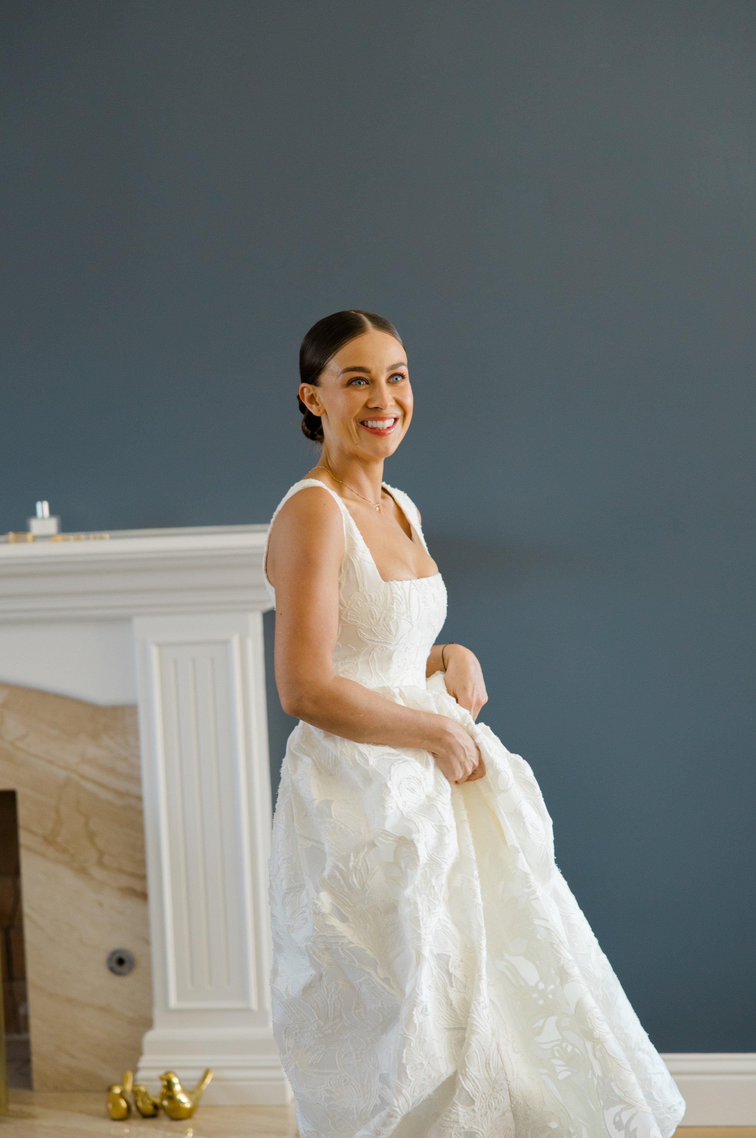 A woman in a white wedding dress standing indoors, smiling, with a blue wall and a white fireplace mantel behind her.