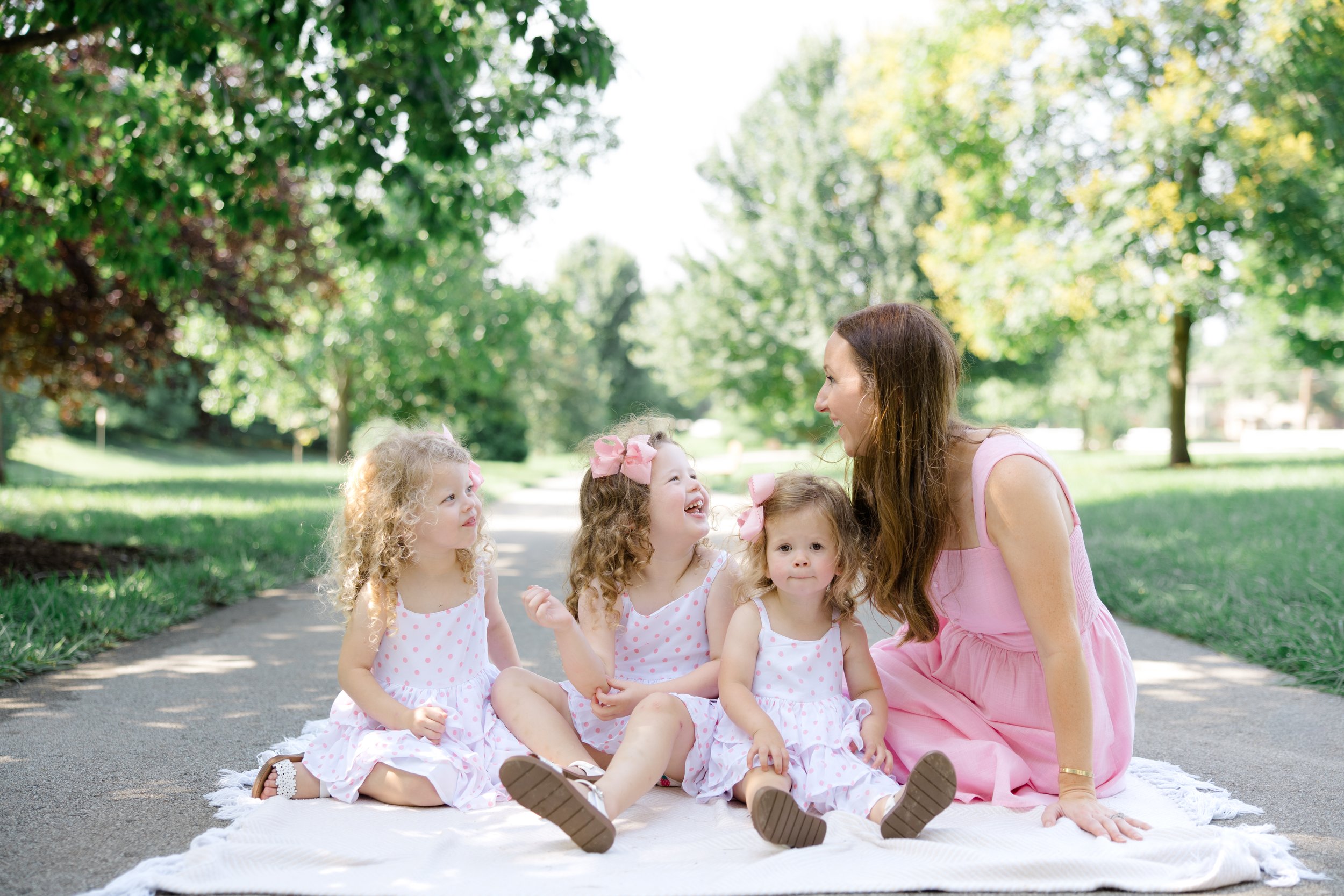 A woman and five young girls sitting on a blanket outdoors in a park surrounded by green trees, smiling and talking with each other.