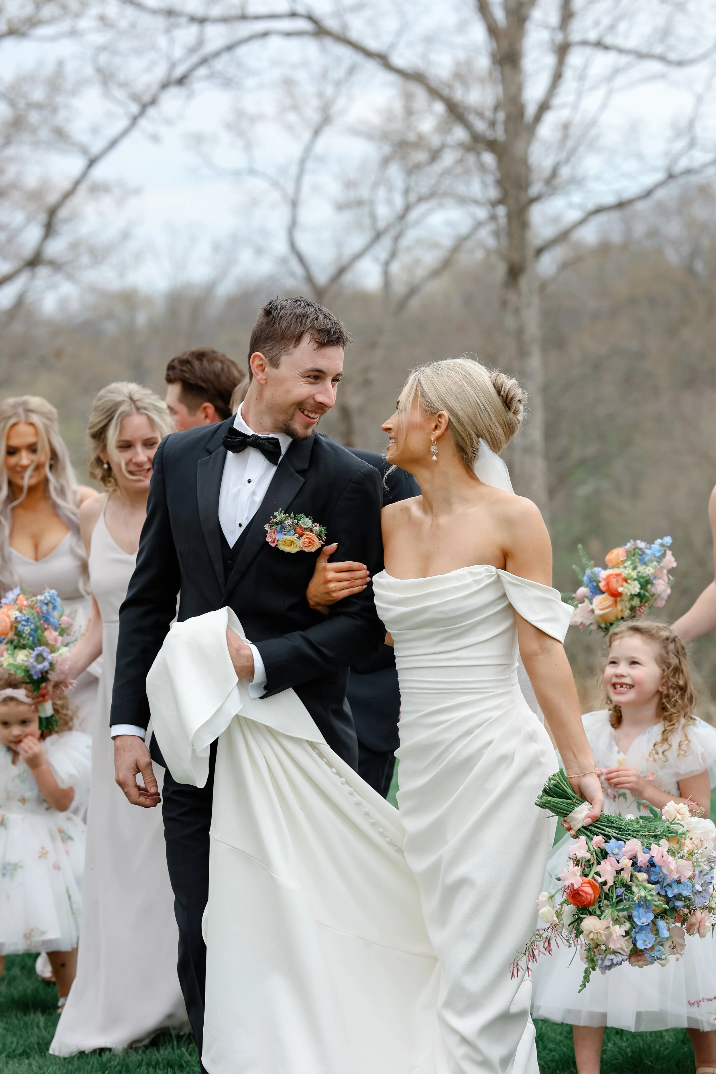 A wedding party outdoors with a bride and groom in the center, smiling and looking at each other, surrounded by women and children holding flowers
